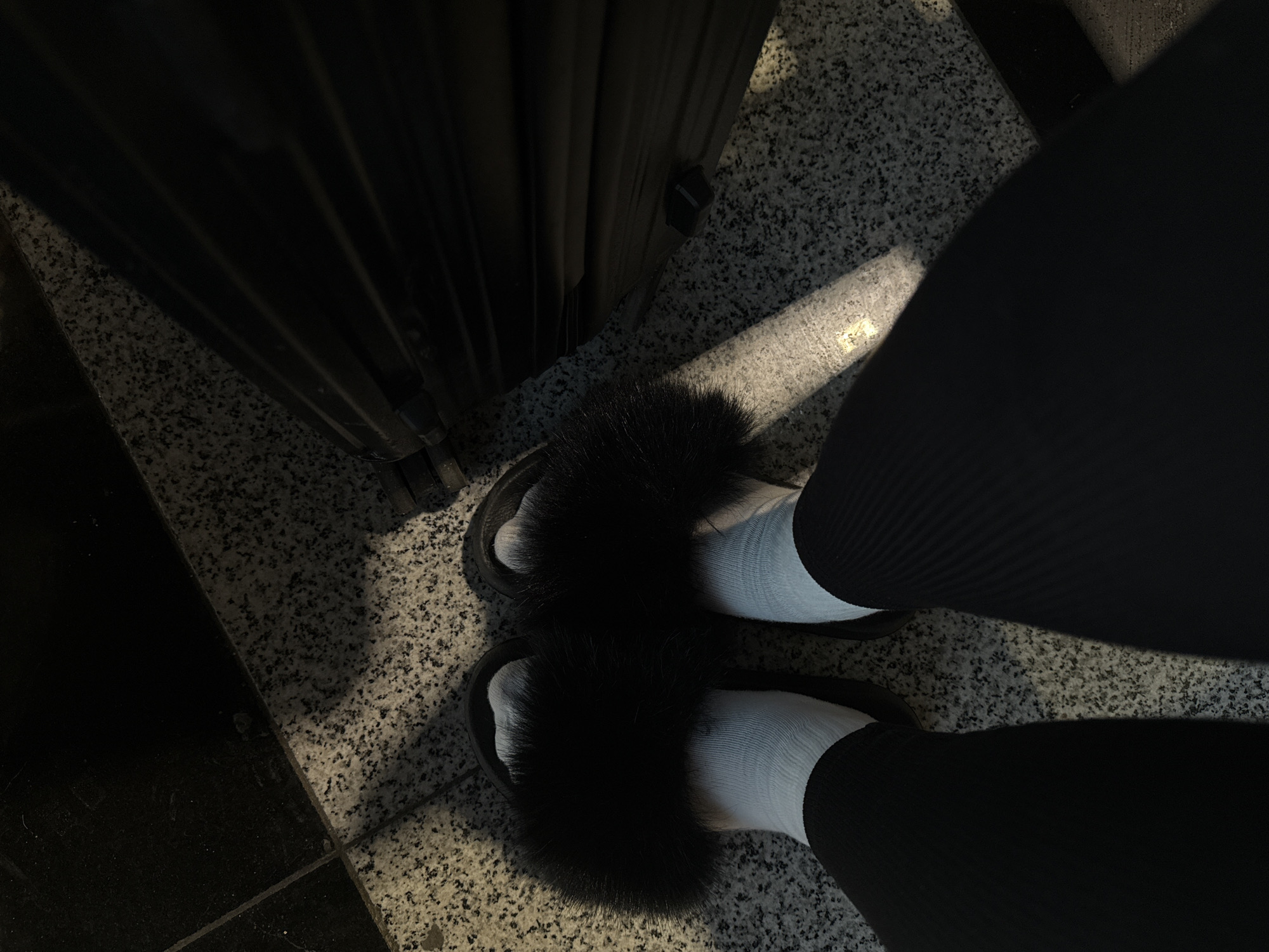 The image, taken in Khimki, Russia, provides a high-angle, close-up view of a person's lower legs and feet in an indoor setting. The individual wears black ribbed pants or leggings, white crew socks, and black, fluffy slide sandals. Their feet are positioned on a grey, speckled floor, resembling granite or terrazzo.
To the left of the frame, a large, dark, vertical object with ribbed detailing is visible, possibly a piece of luggage, a curtain, or a room divider. A strong, diagonal beam of light illuminates a section of the speckled floor and parts of the footwear, casting distinct shadows and highlighting the texture of the floor. In the bottom left corner, a portion of a darker, possibly black, floor tile is seen, contrasting with the lighter, speckled surface. The scene suggests a transient location such as a hotel hallway, lobby, or airport. No specific activity beyond standing is discernible, and no visible text is present. The prominent lighting creates a stark contrast within the frame.