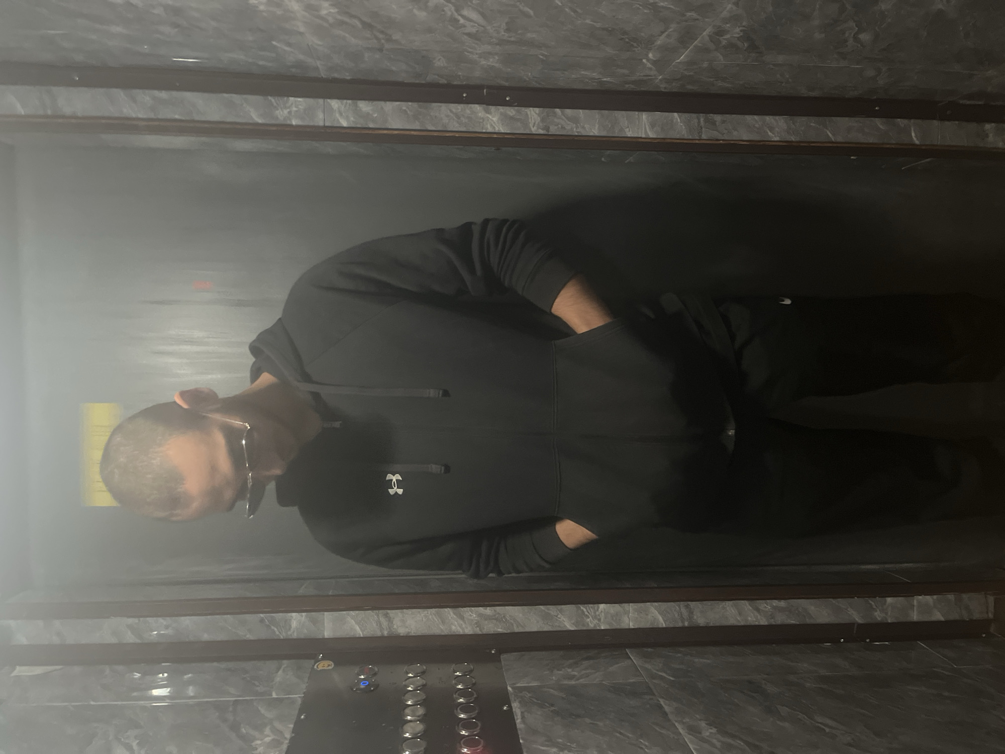 The image, taken in Varna, Bulgaria, shows a person standing inside what appears to be an elevator or a small enclosed space. The photograph is oriented vertically, with the person's head to the left and feet to the right of the frame.
The central subject is a man with short, dark hair, wearing glasses. He is dressed in a black hooded sweatshirt featuring a white "Under Armour" logo prominently displayed on the chest, and black pants. His hands are casually tucked into the pockets of his sweatshirt, and he is looking downwards.
The interior of the space is characterized by dark gray marbled tiles covering both the walls and what appears to be the floor. There are dark metallic or wooden trim elements horizontally dividing the tiled sections. On the lower right side of the image (when correctly oriented, it would be the bottom right of the elevator cab), an elevator control panel with numerous silver buttons is visible. Some buttons are illuminated, showing blue and red lights. The lighting in the space is dim, with a noticeable hazy or foggy effect, particularly on the left side of the frame, possibly from a light source or reflection. No specific activity is taking place beyond the person standing within the enclosed area. The overall impression is one of a modern, utilitarian indoor setting.