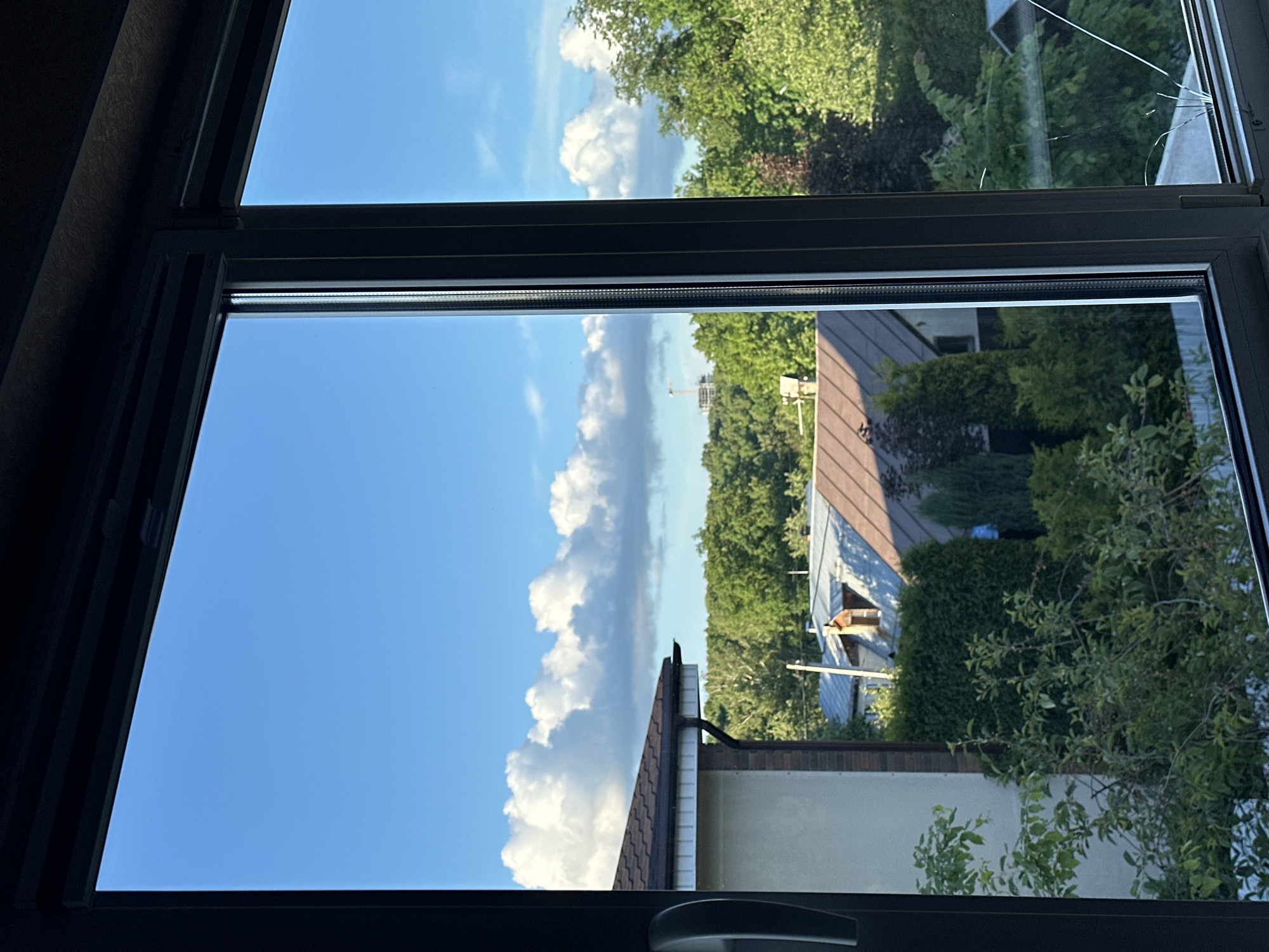 The image shows a view from a window, likely in a residential building. The window is framed by a dark gray or black frame. The upper right pane of the window is cracked.
Outside, the scene includes:
- A blue sky with puffy white clouds.
- Green trees and foliage.
- Rooftops of nearby houses. Some roofs are dark brown, others are light gray.
- A light-colored wall of a building is visible.
- A chimney is visible on one of the roofs.
Given the general appearance of the architecture and vegetation, the scene seems to be in a suburban or residential area. There is no strong evidence in the picture to confirm that the location is in Nizhny Novgorod, Russia. The description does not include any people, actions, or interactions.