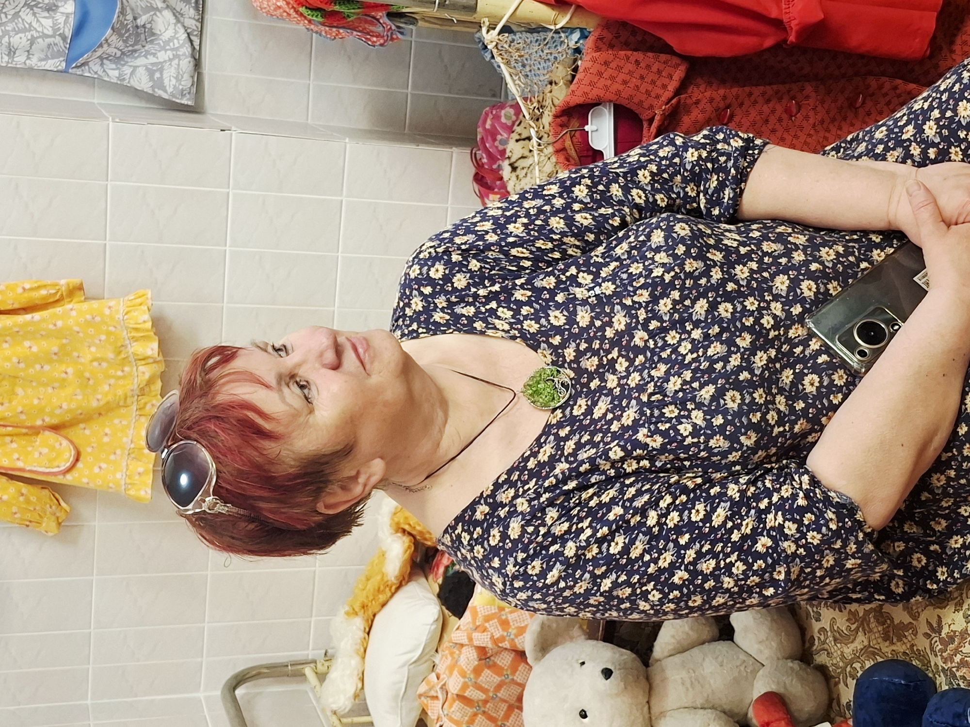 The image features a woman with short red hair wearing sunglasses resting on her head. She is wearing a navy blue dress with a floral pattern and a necklace with a green pendant. She is holding a smartphone in her hands.
The woman is positioned among several objects, including a yellow dress with a floral pattern, various stuffed animals (including a teddy bear), and some red clothing. The background consists of tiled walls.
Based on the location metadata, the scene is in Марковский, Russia.