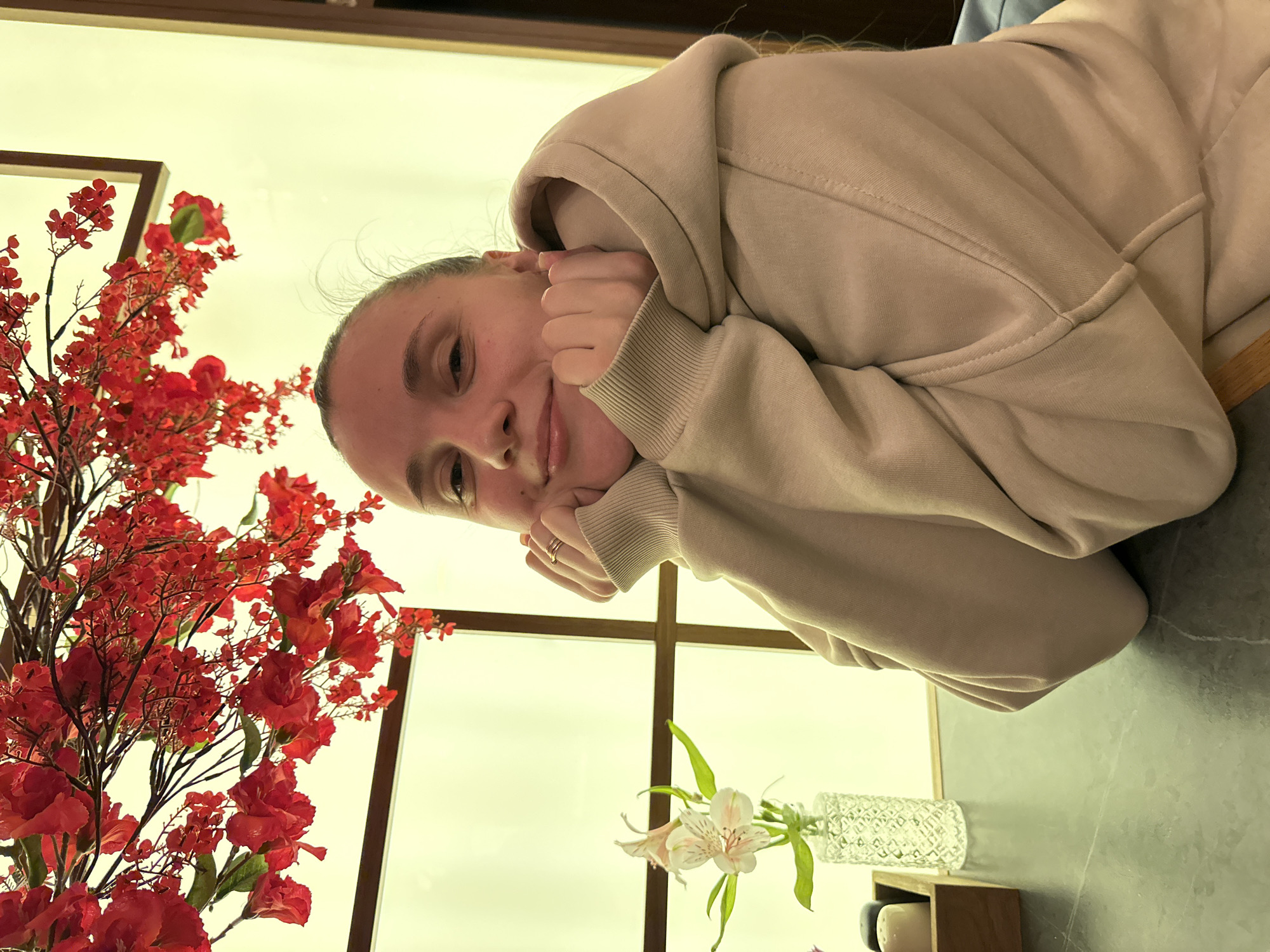 A woman with fair skin and her hair pulled back is reclining, her face resting on her hands, in a room with a cream-colored wall. She is wearing a tan-colored hoodie. Next to her is a large vase of red flowers on the left and a small vase with white flowers to the lower right. The scene is indoors. The location is Rostov-on-Don, Russia.