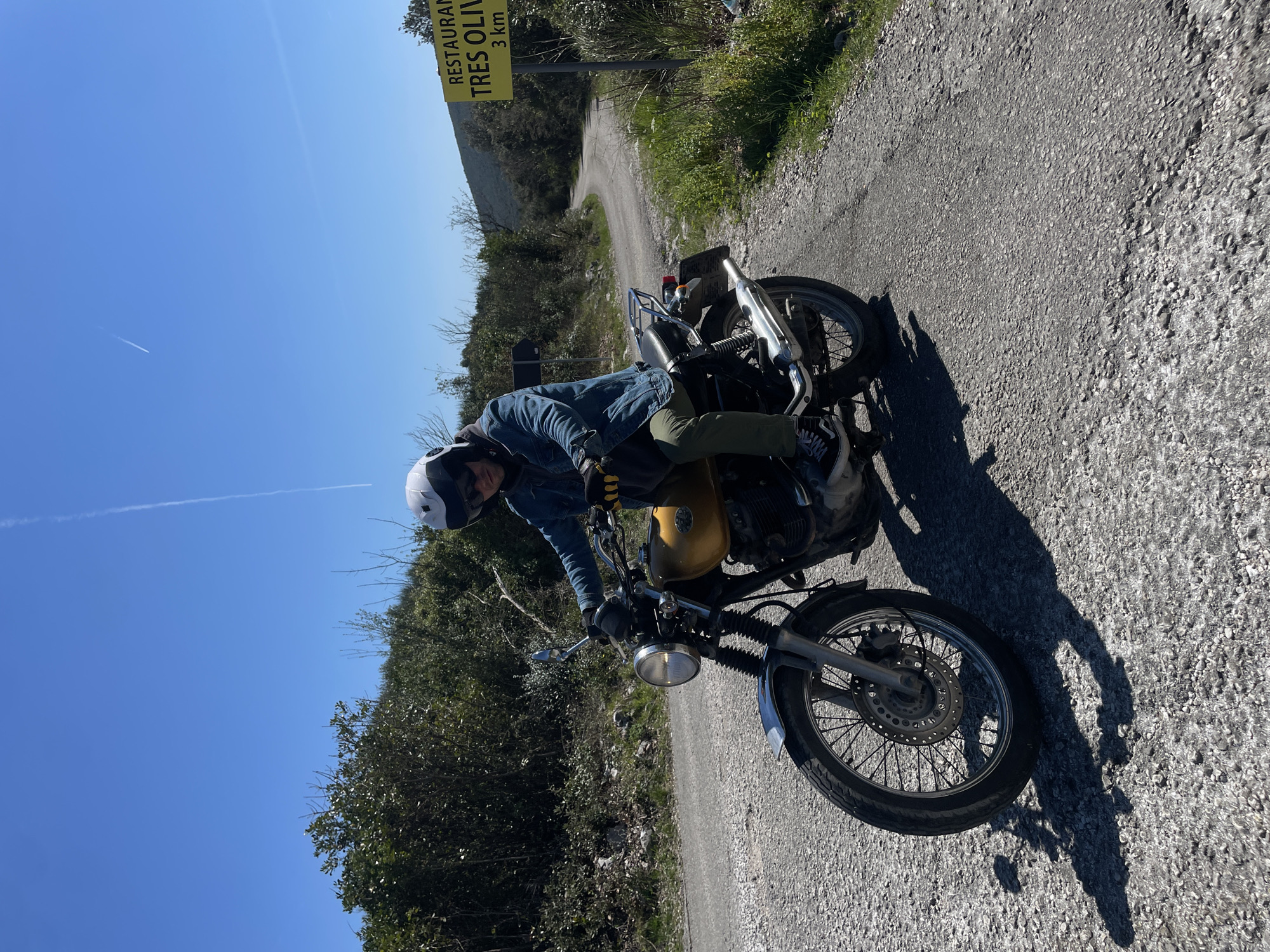 A person wearing a white helmet, blue jacket, and green pants is riding a gold-colored motorcycle on a narrow, winding road in a mountainous area. The road is gray and gravelly. On the side of the road is vegetation, including shrubs and grass. A yellow sign reading "RESTAURANTE TRES OLIVOS 3 km" is visible. The sky is clear and blue. The location is Kubasi, Montenegro.