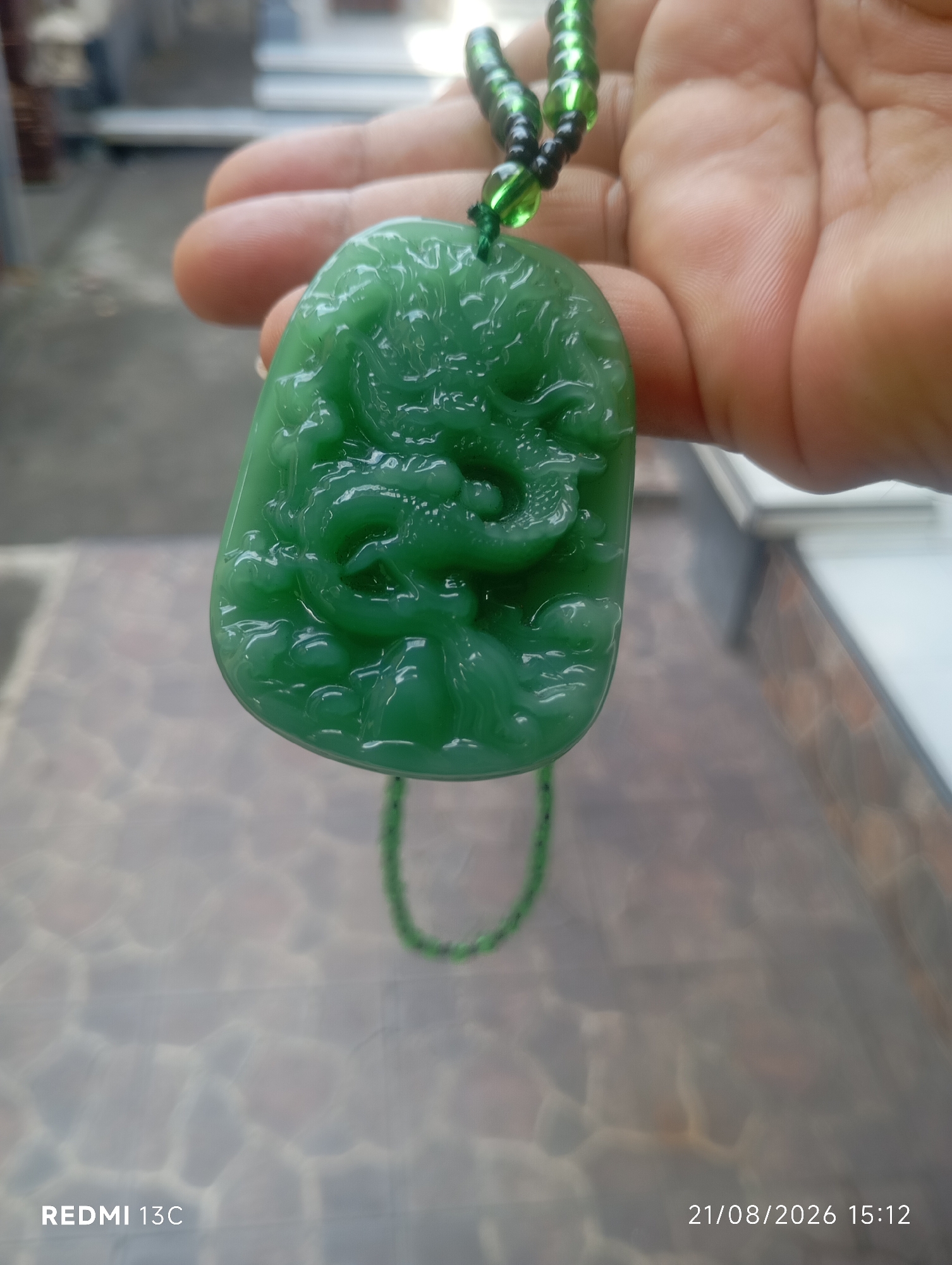 An overhead medium shot shows a person's hand holding a green jade pendant carved with a dragon design. The pendant is strung on a beaded necklace of green and black beads. The background is blurred but appears to be an outdoor setting with a tiled floor and some architectural elements. A timestamp in the lower right corner reads "21/08/2026 15:12". The text "REDMI 13C" is visible in the lower left corner. This image was likely taken in Buleleng, Indonesia.