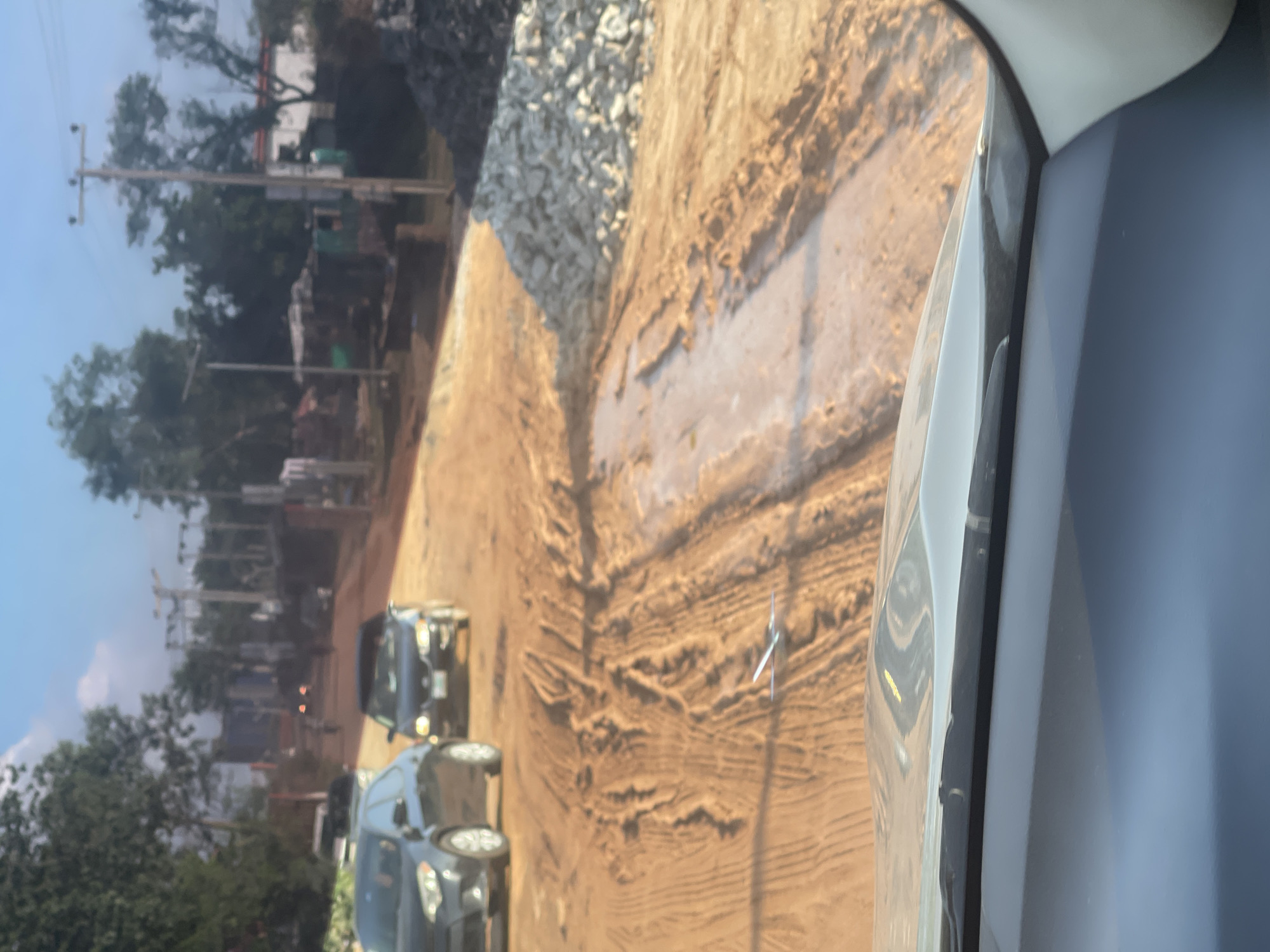 The image, taken from inside a vehicle, shows a road construction site in Abuja, Nigeria. The road is unpaved and has exposed layers of earth. Along the side of the road, there is a retaining wall made of large rocks. Two dark-colored cars are visible on the road. In the background, there are trees, buildings, and utility poles.