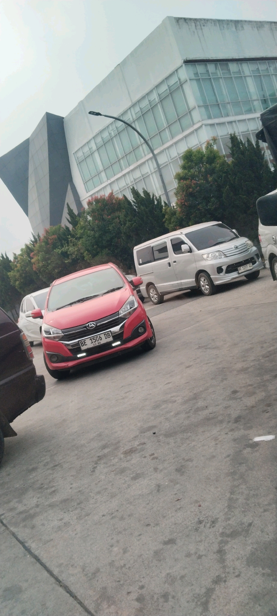 A red Daihatsu Ayla car is parked in front of a modern building with a glass facade and geometric architectural elements. The car's license plate reads "BE 1506 DB." To the right of the Ayla, a silver Daihatsu Luxio van is parked, with its license plate reading "BE 1197 GI." In the background, a row of trees with red and green foliage is visible. The ground is paved with concrete. The sky is overcast. The scene appears to be a parking lot in an urban or suburban area. The location is identified as Haduyang, Indonesia.