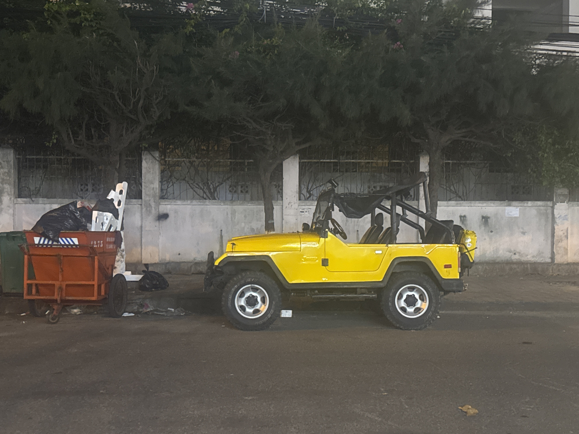 In Bac Nha Trang Ward, Vietnam, a yellow Jeep Wrangler sits parked on a paved street next to a row of trees. The Jeep has its top down and appears to be in good condition. To the left of the Jeep, an orange trash cart is overflowing with black garbage bags and other refuse. A green trash bin is partially visible behind the orange cart. The street is lined with a concrete wall and a decorative metal fence, with trees and foliage growing behind it. The scene is lit by artificial lights, suggesting it is nighttime or early morning. The overall impression is one of an urban street scene with a focus on the contrast between the bright yellow Jeep and the surrounding elements.