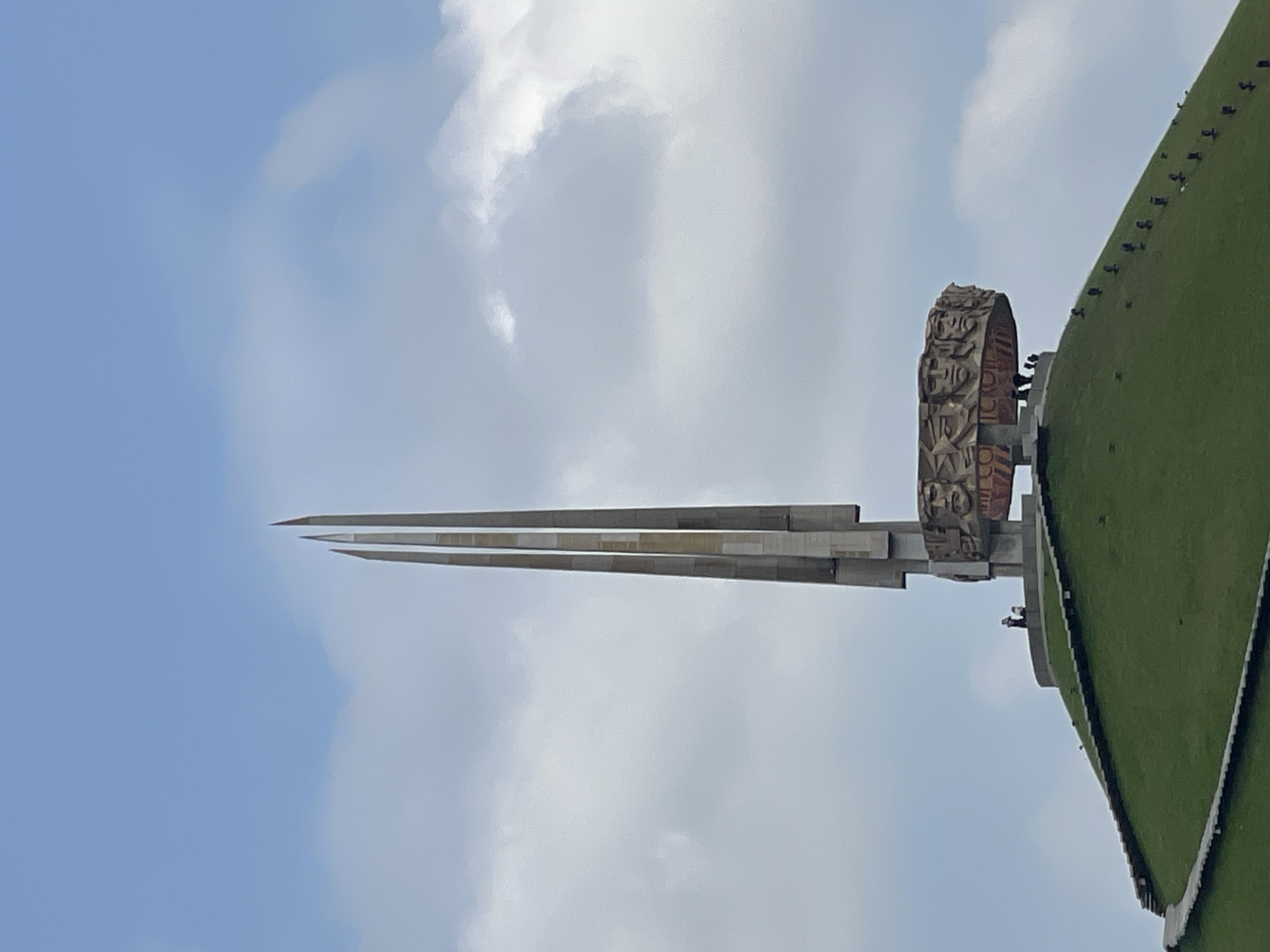 This is a vertical, medium shot of the Victory Monument in the Ozyaritska-Slabadski rural council, Belarus. The monument is a tall, sharp, needle-like structure made of concrete and metal, with three distinct triangular levels converging at a point. To the right of the needle, there's a large, circular sculpture with intricate carvings and what appears to be stylized figures. This circular sculpture is mounted on a concrete base. Beyond the monument, a green, sloped hill rises, covered in grass and with a dark line running along its edge, possibly indicating a path or structure. There are several small, dark figures scattered on the green hill and near the base of the circular sculpture, which are likely people. The sky is a pale blue with scattered white clouds, suggesting it is daytime. The overall impression is one of grandeur and memorial.