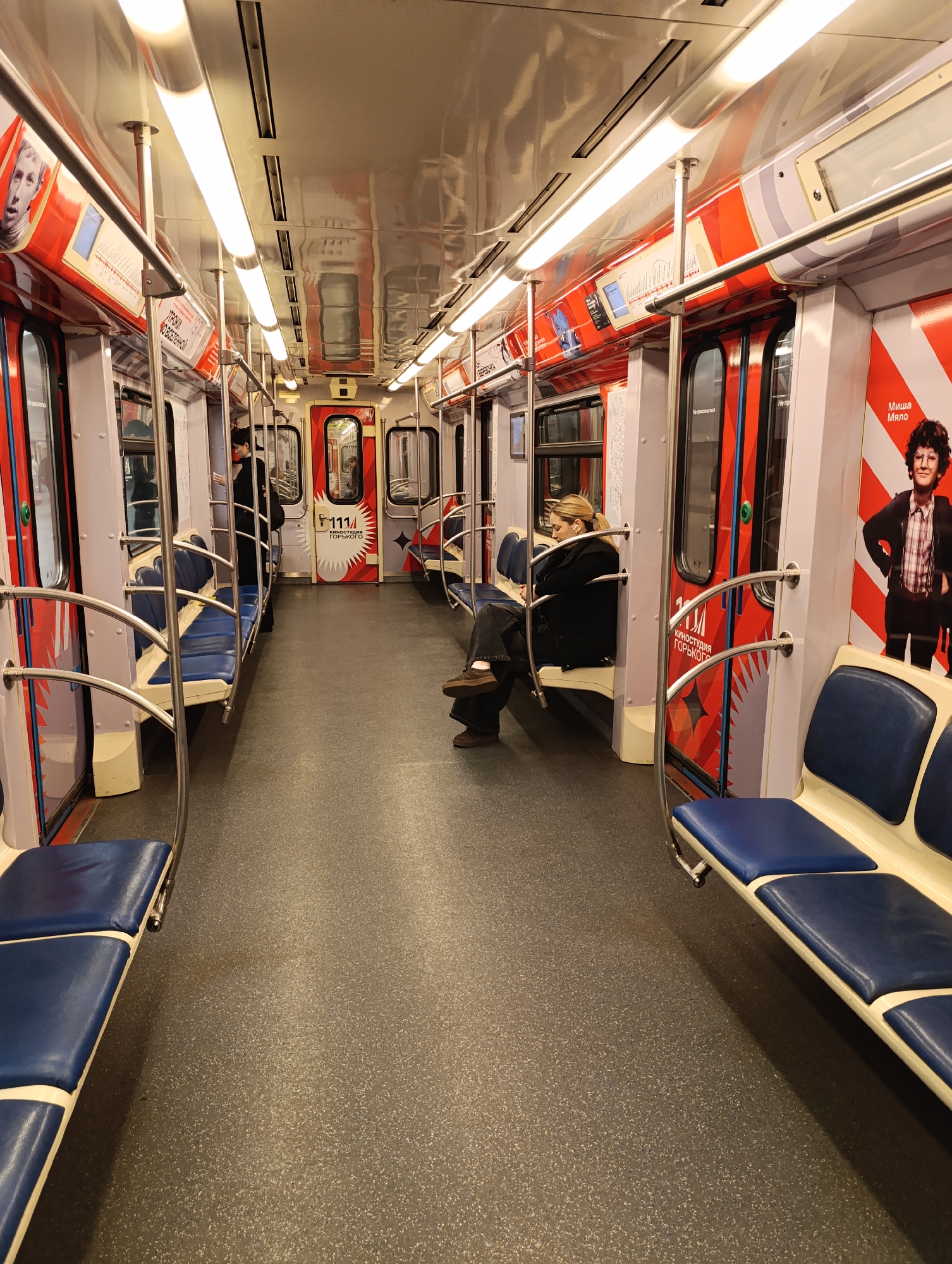 The image depicts the interior of a modern subway car, appearing well-maintained and brightly lit. The scene is set inside a moving or stationary train, characterized by its linear layout, multiple doors, and rows of seating.
The car's interior features a gray speckled floor, blue upholstered bench seats, and white panels. The upper walls are adorned with a distinctive design of red, white, and light gray stripes and patterns. Chrome handrails and vertical poles are strategically placed for passenger support, running along the ceiling and beside the seats. Overhead, a series of bright fluorescent light tubes illuminate the cabin, reflecting off the polished ceiling.
Several individuals are present. In the mid-ground, a woman with blonde hair in a ponytail sits alone, focused on what appears to be a phone or book in her lap. She is dressed in dark clothing, including a jacket and trousers. Further down the car, another person in a dark coat is visible standing near a door. To the right, only the lower legs and feet of a person in brown shoes are visible, suggesting another seated passenger out of frame. The general activity is people riding public transport.
Notable details include the specific aesthetic of the car, which incorporates advertisements and graphics. Prominently visible Cyrillic text provides location cues. On a red panel, the text "Миша Мяло" (Misha Myalo) accompanies an image of a man. Another red panel, styled like a door, features "111 КИНОСТУДИЯ ГОРЬКОГО" (111 GORKY FILM STUDIO). Smaller, cautionary text on the actual doors reads "Не прислоняться" (Do not lean). Additional Cyrillic text on the upper wall decor includes "УРОКИ ВСЕОБЩЕЙ" (Lessons of Universal/General) and "ВЫСТАВКИ" (Exhibitions), suggesting informational or promotional content. The interior lighting indicates it's an indoor setting, and the time of day is not discernible from the image.