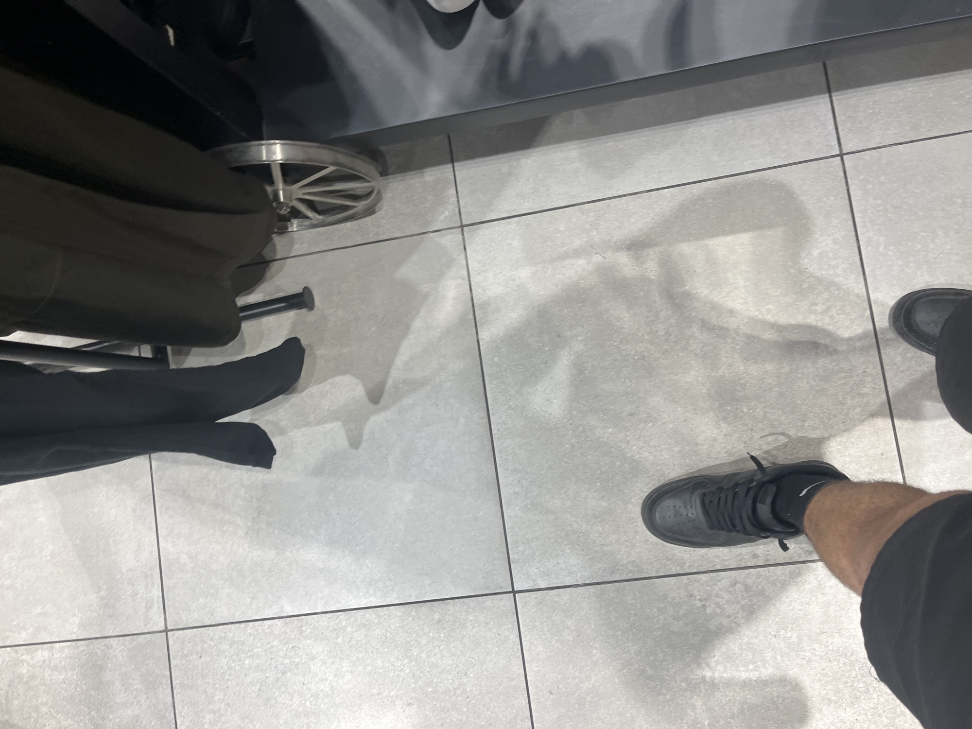 A high-angle shot shows a person's legs and shoes standing on a light grey tiled floor. The legs are clad in black clothing. One leg is visible from the knee down, wearing black sneakers with black socks. The other leg is less visible, with only a black shoe and the lower part of the pants seen. The tiles are rectangular, and dark grout lines separate them. In the upper-left portion of the image, there is a black chair or piece of furniture with metal legs and wheels. The image captures shadows, suggesting overhead lighting. The location of the photo is indeterminable as there are no identifying markers that are particular to the city of Blagoevgrad, Bulgaria.