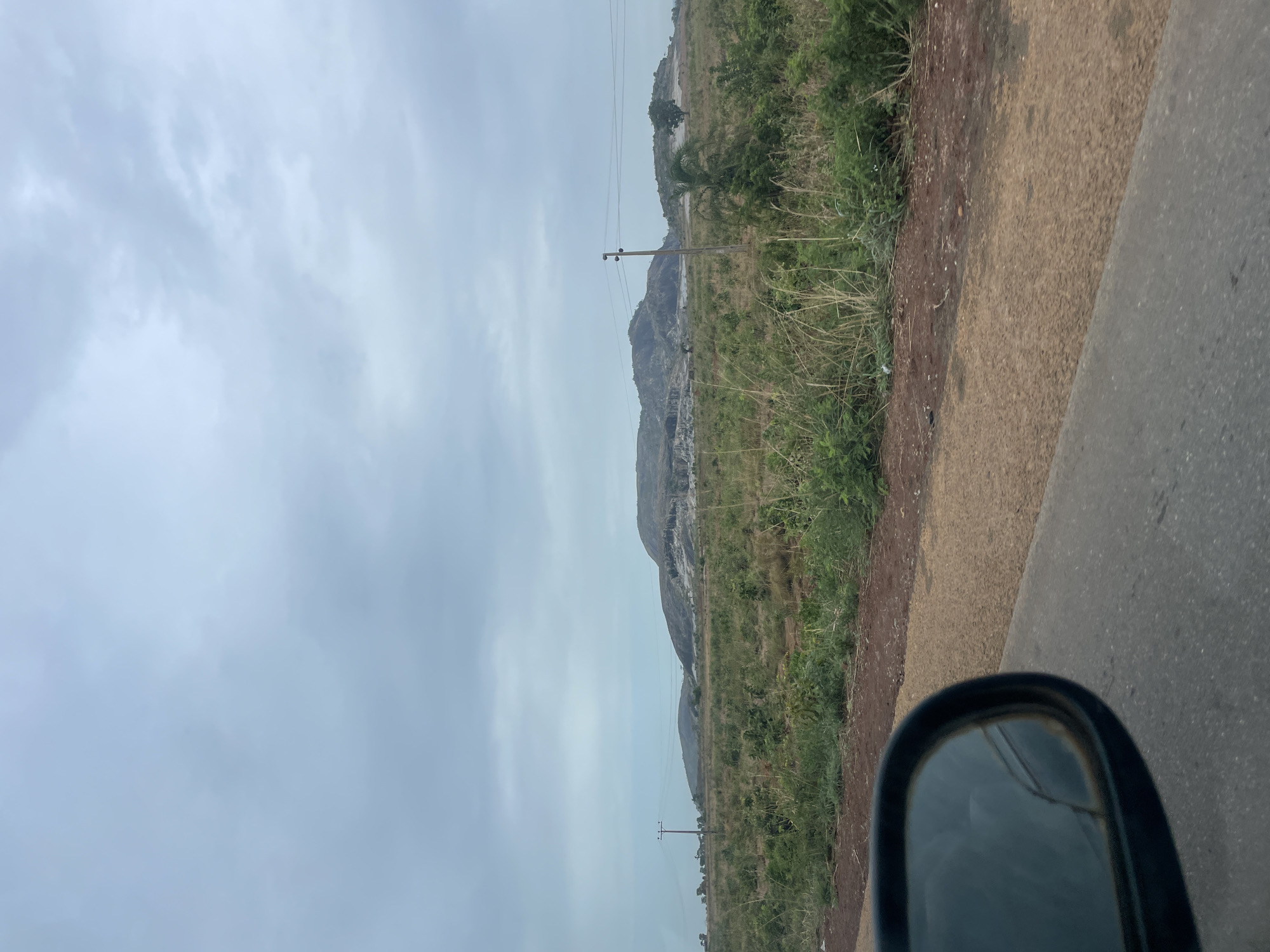 The image shows a landscape in Kujama, Nigeria, viewed from what appears to be the interior of a vehicle. A cloudy sky dominates the left portion of the image. In the center, there's a view of distant, grassy terrain leading to a rocky hill or small mountain. There is a utility pole with power lines. To the right, a narrow strip of roadside dirt and vegetation is adjacent to a paved road surface. In the bottom right corner, a side mirror of the vehicle is visible.