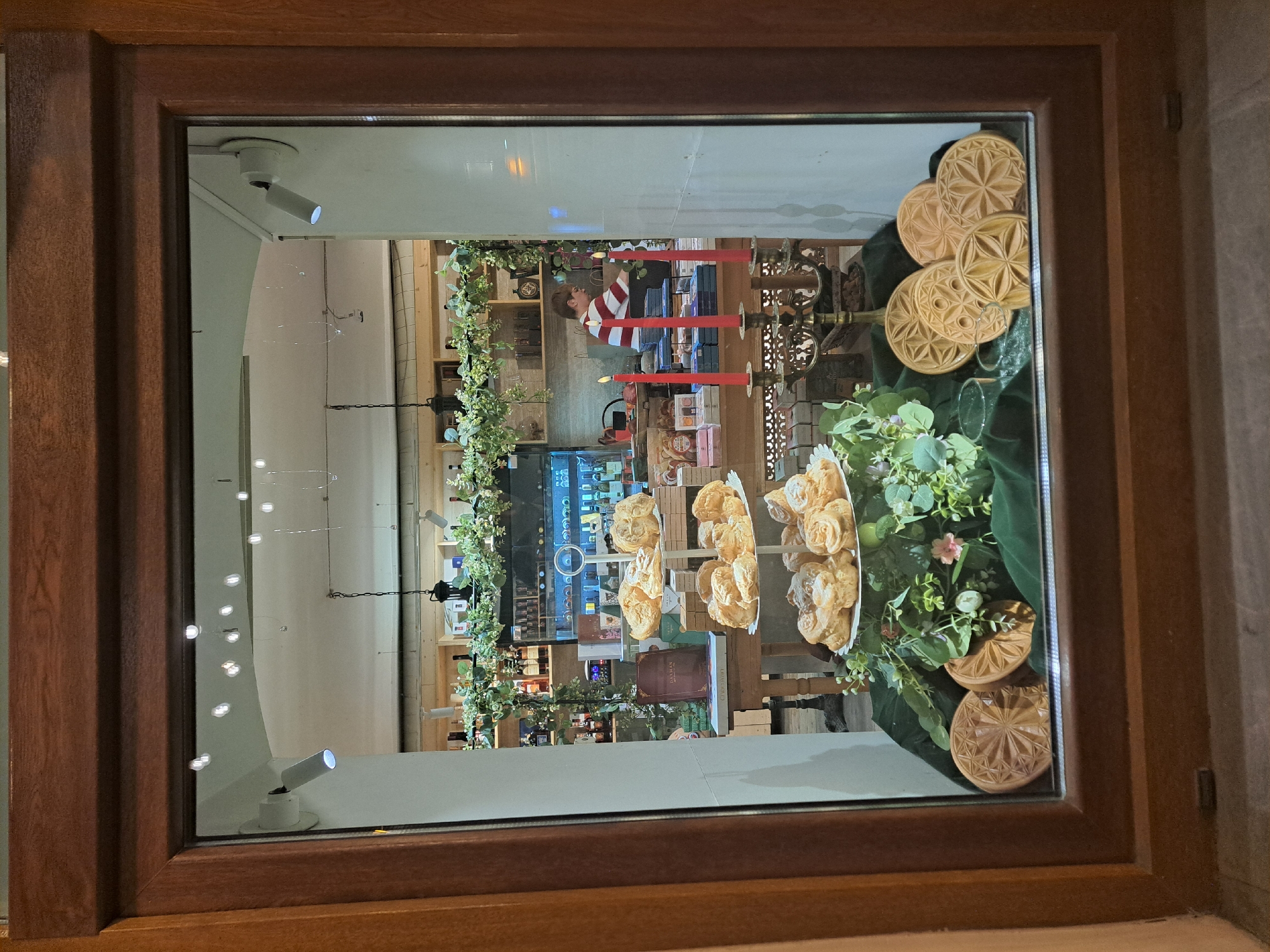This is an interior scene photographed through a window. The location is likely in Yerevan, Armenia given the context. Inside, a person wearing a striped red and white shirt is partially visible in the background, standing behind a counter. There are decorative elements, including garlands with lights and green foliage, along with red candles. Baked goods, possibly pastries or cookies, are displayed on stands and platters. Wooden carved round objects are also arranged. The store seems to sell or display food products and decorative items. The environment suggests a cafe or retail shop with a holiday or special occasion theme. The reflection on the glass and the angle of the photo make it harder to determine everything that is going on within the scene. There are lights mounted on the ceiling.
