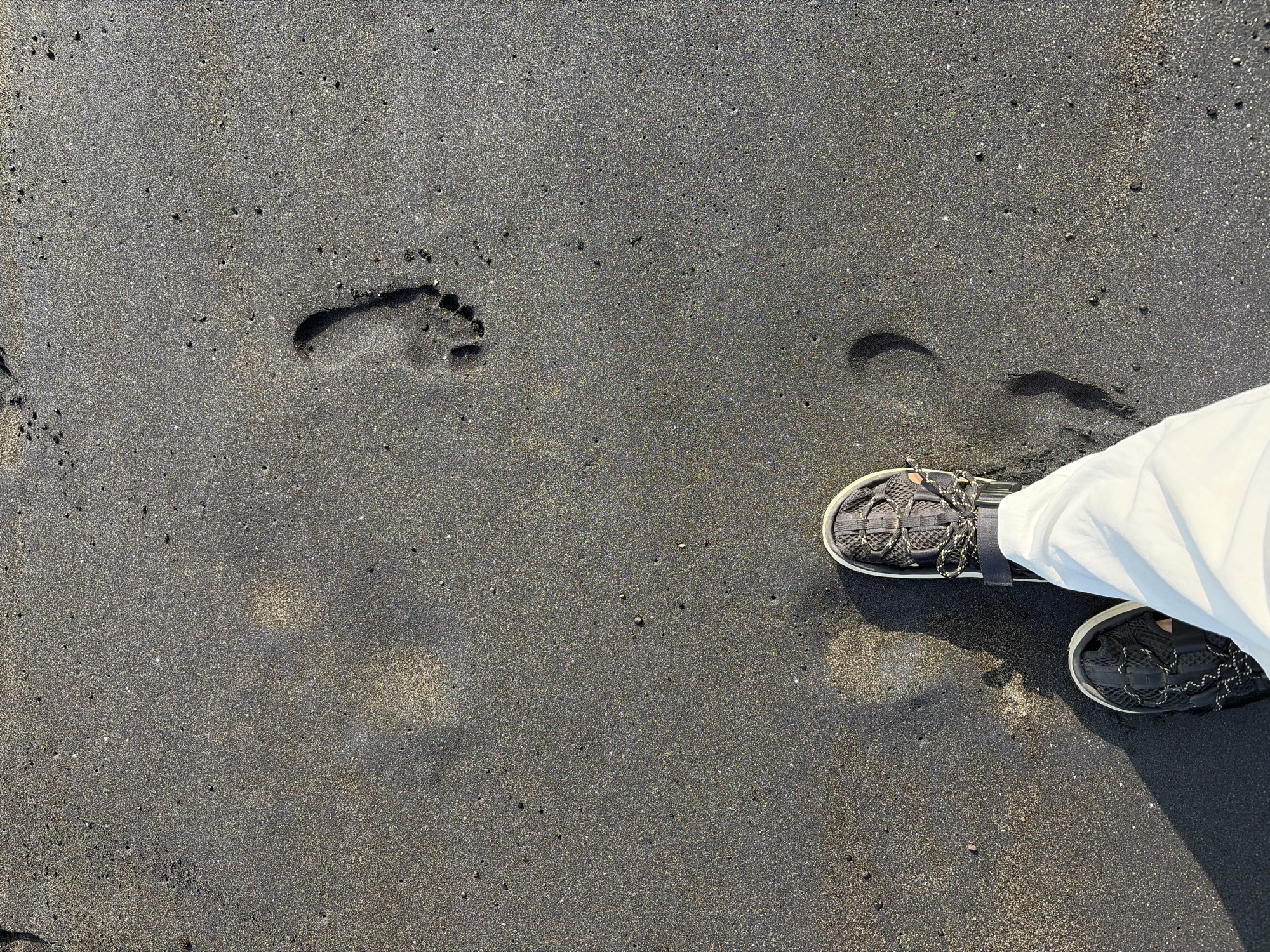 The image shows a close-up, overhead view of a person's feet in dark sand on a beach. The sand is a fine-grained, dark volcanic sand, characteristic of some beaches in Bali, Indonesia. There are several footprints visible in the sand, indicating recent passage. The person is wearing dark, casual sandals with white soles and white pants. The sunlight casts shadows on the sand, suggesting it is daytime. The overall scene is one of a leisurely walk on a beach, with the focus on the interaction between the person's feet and the sand. The location is identified as Blahbatuh, Indonesia.