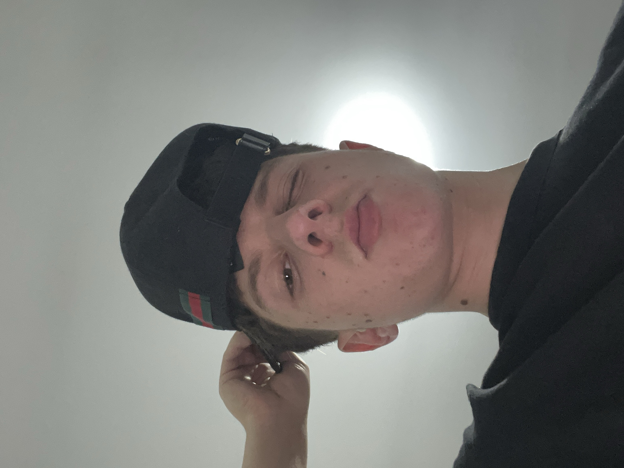 A male individual, appearing to be a young adult, is captured in a low-angle close-up portrait. The scene is situated in what is reported as Zlatograd, Bulgaria. He is wearing a black t-shirt and a black baseball cap featuring a distinctive red and green striped band on the rear. His head is slightly tilted upwards, and he is looking directly into the camera with his left eye open and his right eye winked closed. His lips are pursed in a pouting expression, and several freckles are visible on his nose and cheeks. His left hand is raised, with fingers interacting with the hair near the back of his head, beneath the cap. The background consists of a plain, light-grey or white surface, consistent with a ceiling, which is largely featureless. A bright, diffuse light source is visible in the upper right quadrant of the image, illuminating the background and creating rim lighting on the individual's right jawline and neck.