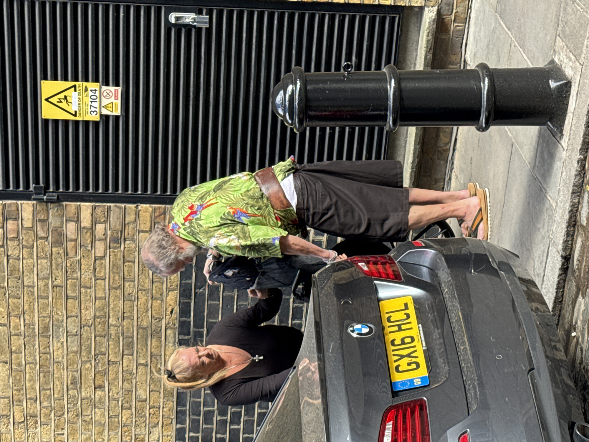 The image shows two individuals, an elderly man and a middle-aged woman, near a dark gray BMW car in what appears to be a street setting. The man is wearing a green, flowered shirt and dark shorts, and has gray hair. He is bent over near the car's rear. The woman is wearing a black top and has blond hair pulled back. She is standing near the car's rear as well. The car has a yellow license plate that reads "GX16 HCL". Behind them, there is a wall constructed of tan-colored bricks on the left, and a metal structure with a yellow "DANGER OF DEATH" sign attached to it. To the upper right, there is a short, black bollard. Given the license plate and the general architectural style, the location is likely somewhere in London, United Kingdom.