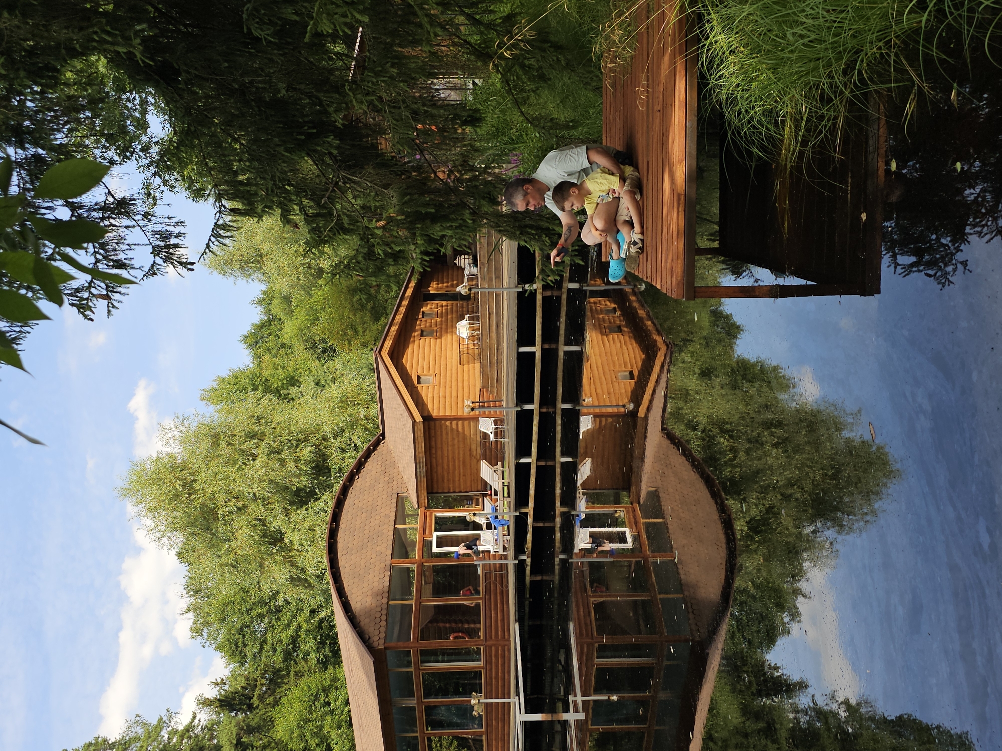 A man and a boy are sitting on a wooden pier on a body of water, with a wooden building with a curved roof in the background. The man, wearing a light-colored t-shirt and pants, is pointing his left index finger towards the water. The boy, wearing a yellow t-shirt and shorts, is sitting next to him, looking in the same direction. They are both seated on the pier with their legs crossed. A blue flotation device is placed near them on the pier. The water reflects the building and trees. The building appears to be a lodge or resort, with large windows and wooden siding. The surrounding environment is lush with green trees and foliage. This scene is located in the village of Ulitkino, Russia.
