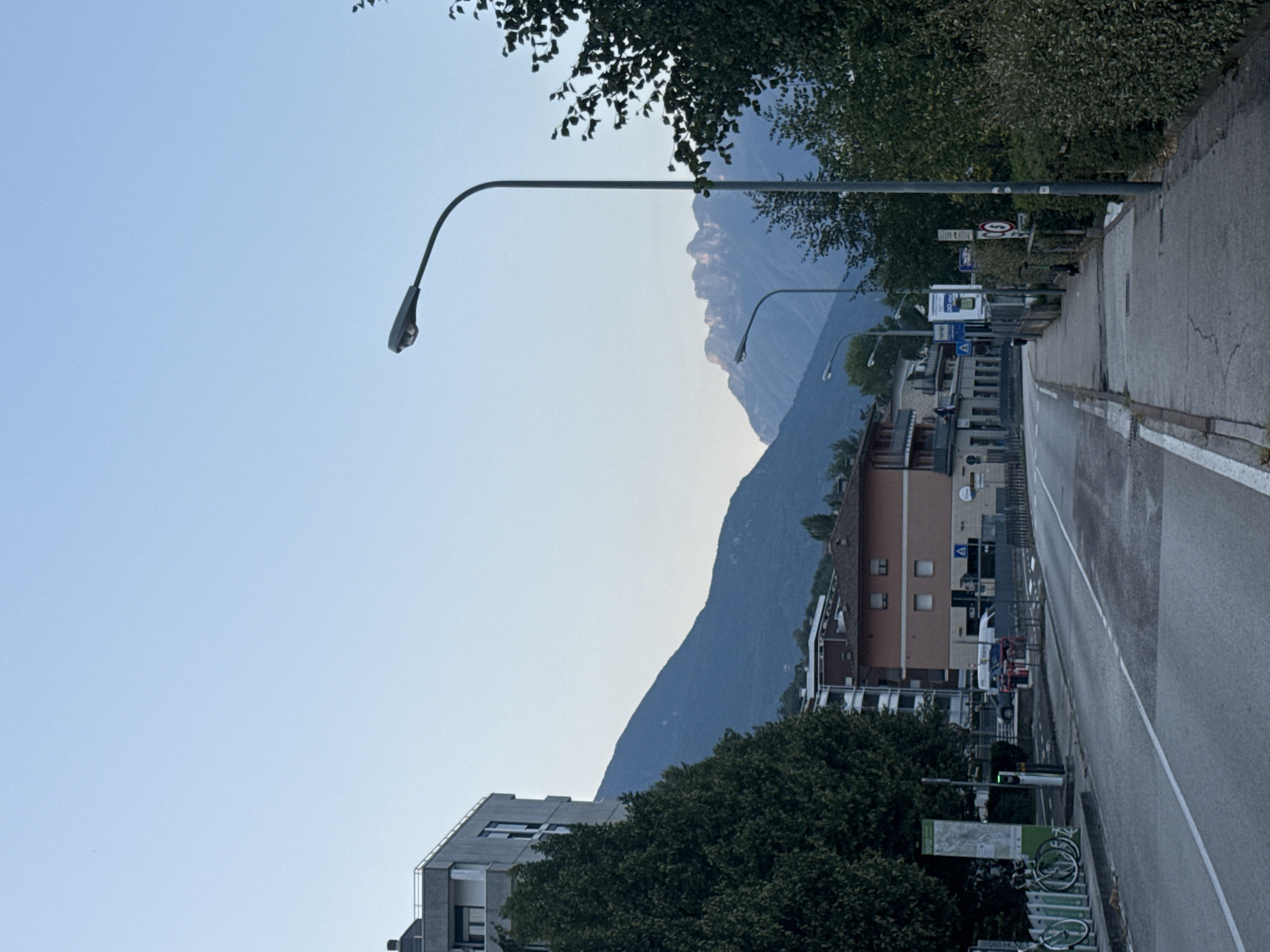 A paved road runs diagonally through the frame, leading away from the viewer towards a range of hazy, blue mountains under a clear, pale blue sky. The road is lined on the left by a sidewalk, a bicycle rack with several bicycles, a tall green information kiosk, and trees. Beyond the trees on the left, a modern, multi-story building with a white railing around its rooftop is visible. On the right side of the road, there is a sidewalk, a tall lamppost, and dense green foliage. Further back, several buildings are visible, including a two-story, reddish-brown building with white-trimmed windows. In the background, the sun appears to be setting or rising, casting a warm glow on the mountain peaks. The overall scene depicts a quiet street in a town nestled among mountains, possibly in the city of Trento, Italy.