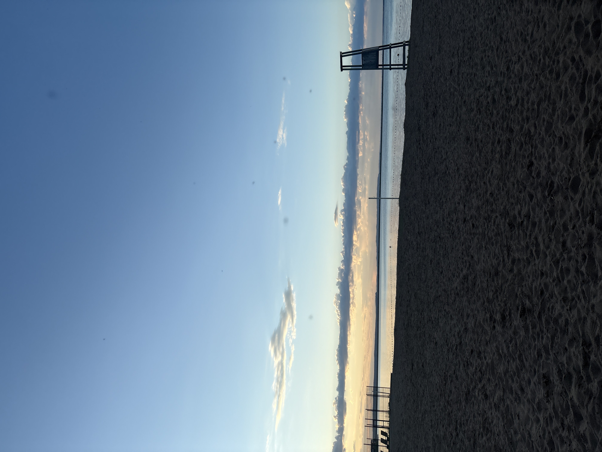 This image depicts a serene beach scene in Chaikovsky, Russia, captured during either sunrise or sunset. The sky dominates the upper portion, displaying a clear blue tone that gradually lightens to golden hues along the horizon, illuminated by the low-angle sun. Several elongated clouds are present, reflecting these warm colors, while a few small, dark specks, likely sensor dust, are noticeable against the blue sky.
The lower half of the image features a wide expanse of dark, textured sand, showing subtle ripples and indentations. To the left, the calm surface of a large body of water, presumably a lake, stretches to meet the horizon. A distant line of small markers or buoys is visible across the water, indicating a designated boundary.
A tall, dark wooden lifeguard chair stands prominently near the water's edge, positioned in the middle-right of the frame. There are no people visible in the scene, contributing to a sense of peaceful solitude. The weather appears clear with scattered clouds. No text is visible in the image.