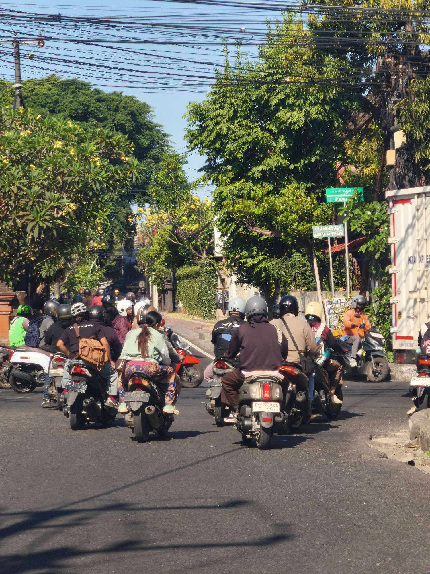 A crowded street in Denpasar, Indonesia, shows a traffic jam of motorcycles. The sky is clear and blue. Sunlight illuminates the scene, casting shadows on the asphalt road. Lush green trees with yellow flowers line the street, creating a vibrant canopy. Buildings with stone walls are visible on the right side of the road. A large white truck is parked on the right. Street signs indicate "JL. KUMBA" and another sign pointing in different directions. Numerous motorcyclists, most wearing helmets, are seen navigating the congested road. Some motorcycles are parked along the side, while others are in motion, contributing to the traffic. The overall atmosphere suggests a typical busy day in a bustling Indonesian city.