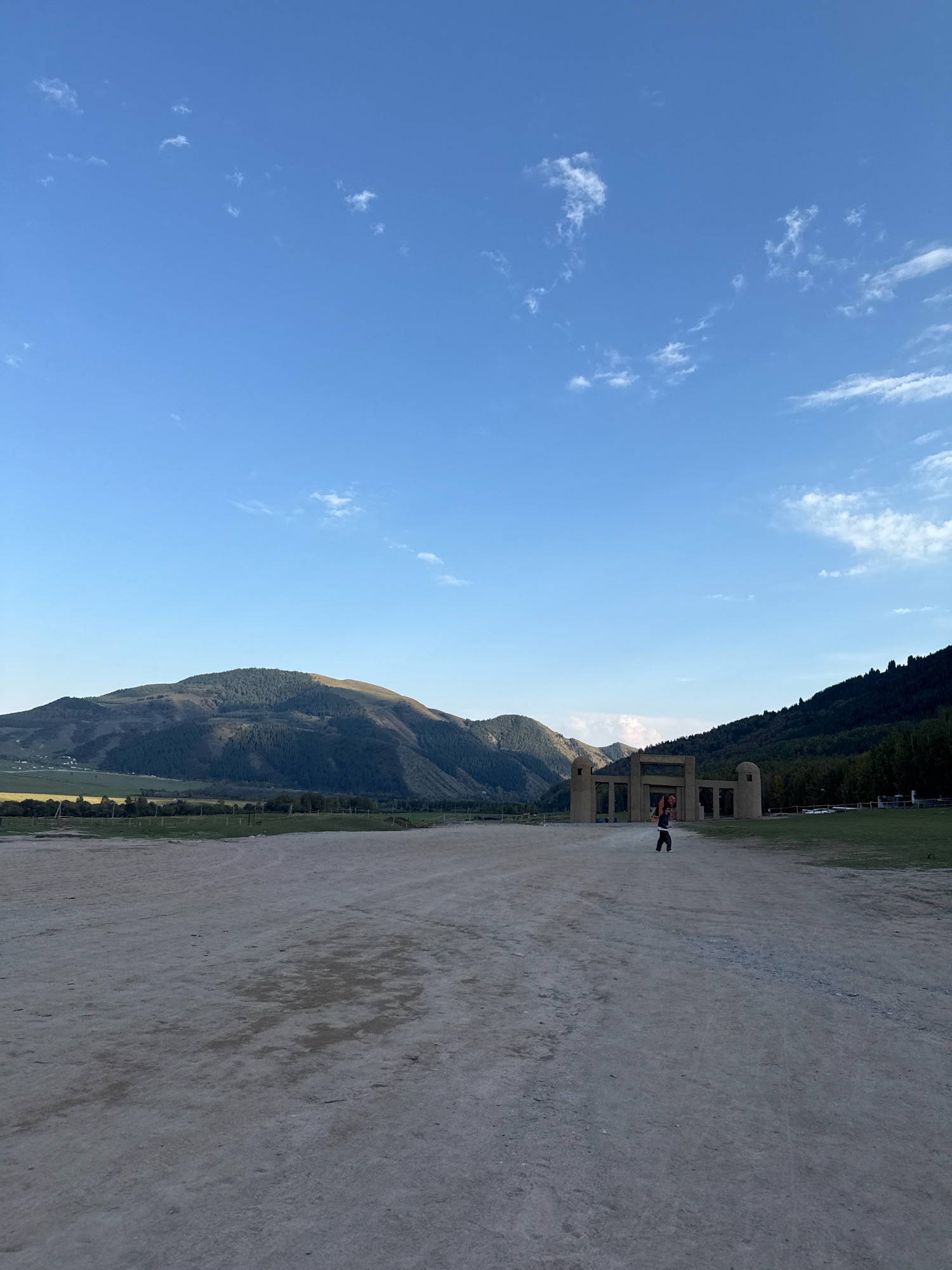 A lone individual walks across a dusty, open expanse toward a large, arched structure, likely an entrance gate, in Semenovka, Kyrgyzstan. The structure, made of tan stone or concrete, features two prominent round turrets flanking a central archway. Beyond the gate, rolling, forested hills rise, with one larger mountain dominating the left side of the frame, its peak catching the sunlight. The sky above is a clear, bright blue with scattered white clouds. The foreground is a wide, dirt path, showing tire tracks and some patchy vegetation. The overall impression is one of a vast, natural landscape with a man-made monument.