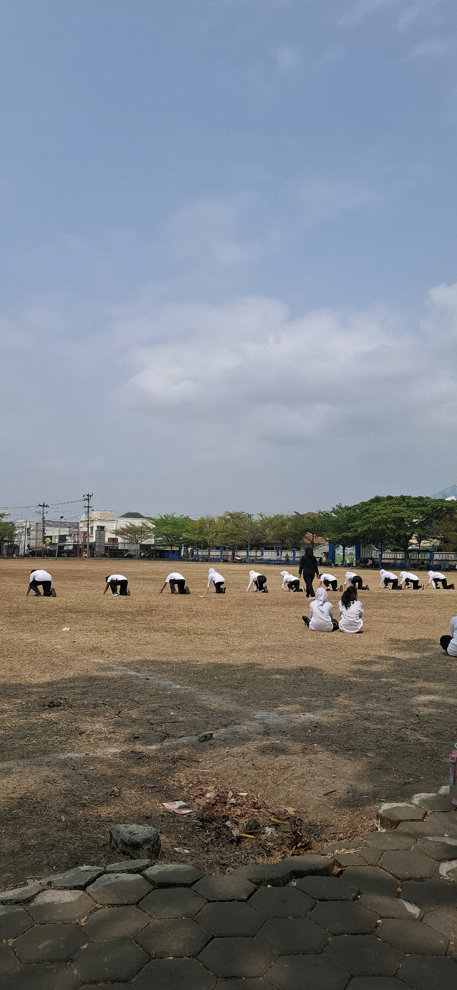 In the city of Baturan, Indonesia, a group of individuals, likely students, are engaged in physical activity on a dry, grassy field under a partially cloudy sky. The scene appears to be an outdoor sports ground or schoolyard. Many of the individuals are wearing white shirts and dark pants or skirts, suggesting a uniform. They are positioned in various stances, some with their knees on the ground and torsos bent forward, while others are seated. One person, dressed in black, is standing and appears to be instructing or overseeing the group. In the background, buildings, trees, and power lines are visible, indicating an urban or semi-urban setting. The ground is covered with dry grass and patches of bare earth, with some debris and paving stones visible in the foreground. The overall impression is one of a structured outdoor exercise session.
