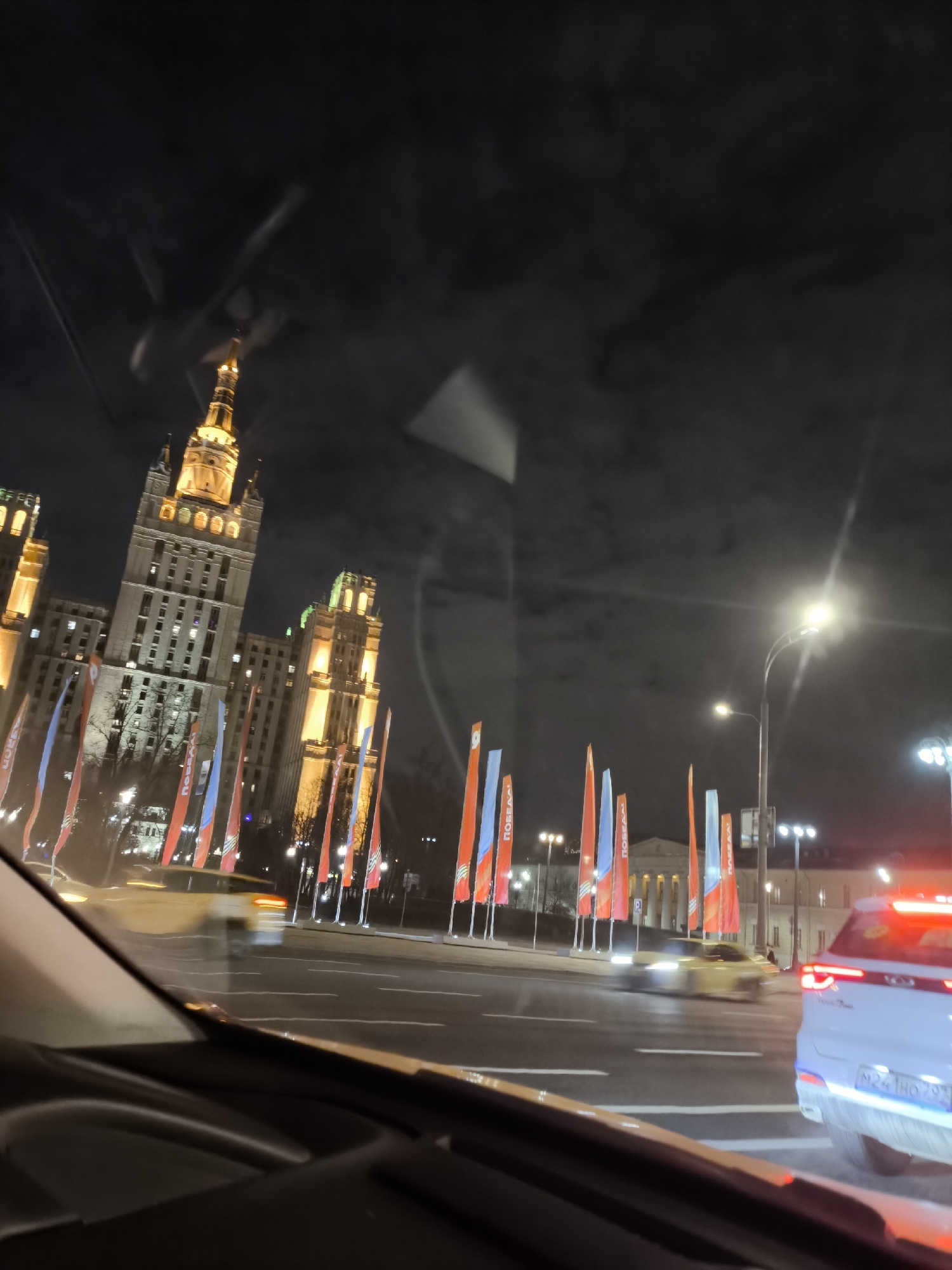 The image displays a nighttime urban scene from the perspective of inside a car, looking out through the windshield. Dominating the left side is a towering, illuminated building, characteristic of Stalinist Empire architecture with a distinctive spire and intricate details, suggestive of one of Moscow's "Seven Sisters." Its facade glows with warm light, and some windows are lit from within.
Along the wide street in the middle ground, numerous flags are displayed. These flags are predominantly red and blue, and several clearly show the Cyrillic word "ПОБЕДА!" (Pobeda!), meaning "Victory!". This indicates a celebratory or commemorative occasion, likely Victory Day.
The road itself is wet or reflective, with lane markings visible. Traffic is present, with a white car in the right foreground displaying a Russian license plate "M241HO 799". Other vehicles, including a yellow car, appear blurred further down the road, suggesting motion. Bright streetlights provide illumination, with some creating lens flares. The sky is dark, confirming the nighttime setting. The bottom portion of the image shows the car's dashboard and reflections on the windshield. The overall scene is consistent with a major thoroughfare in Moscow, Russia.
