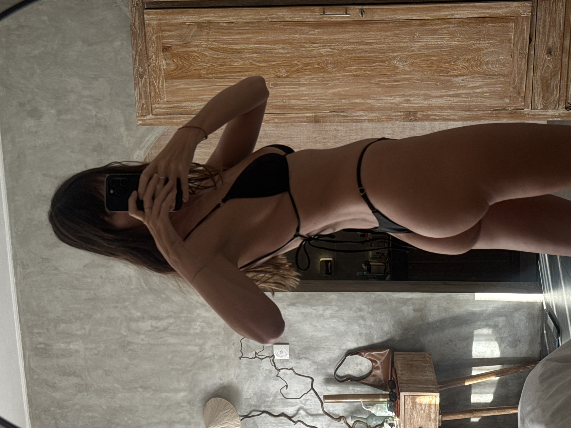 A woman with brown hair wearing a black bikini is taking a photo of herself in a mirror. The background includes a grey textured wall, a wooden cabinet, and what appears to be a shelf with various objects including a brown bag. Sunlight is streaming in from a window. The location is potentially in North Kuta, Indonesia.