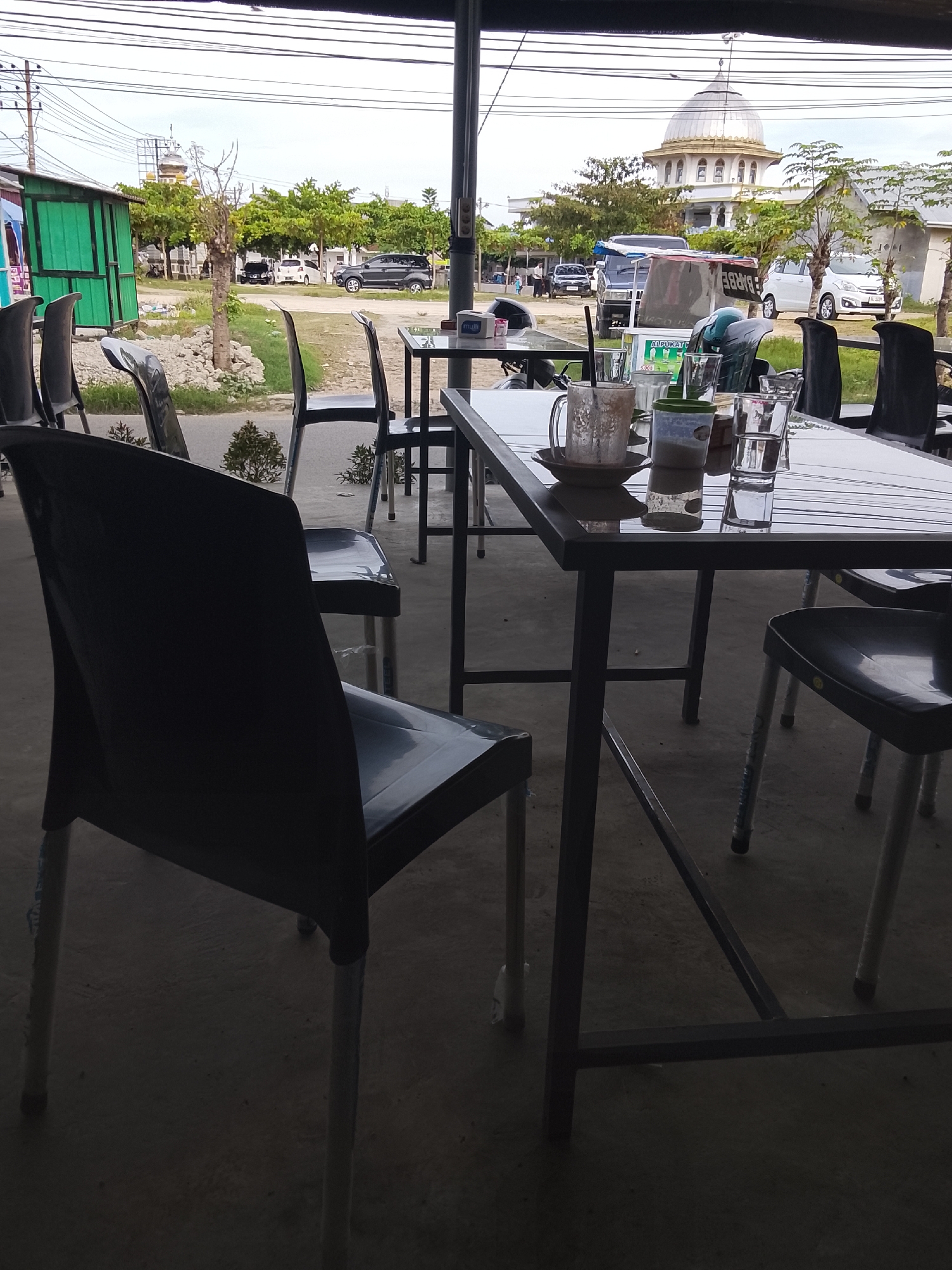 The image shows an outdoor seating area, likely a cafe or restaurant, in Lhokseumawe, Indonesia. Several black plastic chairs with silver metal legs surround tables with reflective surfaces. Used drinkware, including a mug and glasses, is visible on one table. In the background, through the open structure, is a view of a street scene with cars, trees, and a building with a dome resembling a mosque or religious structure. The sky is overcast.