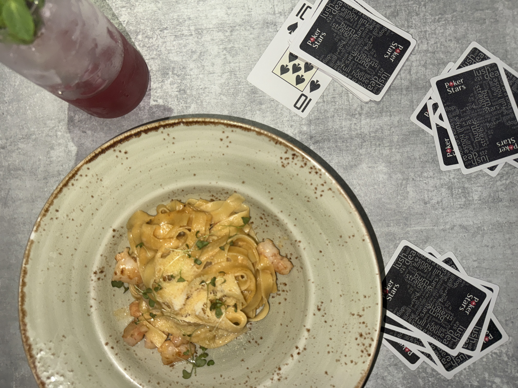 The image displays an overhead view of a light gray, textured table surface. There is no discernible information within the image itself to confirm a location in Perm, Russia.
In the upper-left quadrant, a round, rustic ceramic plate with a speckled, light beige/green finish holds a serving of tagliatelle pasta. The pasta is coated in a light-colored creamy sauce and mixed with cooked shrimp. The dish is garnished with small sprigs of fresh green herbs, possibly microgreens. A distinct white, possibly cheesy or creamy, topping is visible over a portion of the pasta.
To the right of the plate, a tall, clear glass contains a red or pink beverage with ice cubes and a green garnish, likely mint leaves, at the rim. Condensation is evident on the exterior of the glass.
Adjacent to the glass, two standard playing cards are partially visible face-up. One card is clearly identifiable as the 10 of Spades. The other shows a spade suit symbol and is partially obscured.
Across the right and bottom portions of the table, several stacks of face-down playing cards are scattered. The card backs are black with white text, featuring the "Poker Stars" logo in red and white. The background of the card backs is densely covered with repeated poker-related terms such as "lush," "deal," "full house," "burn," "flop," "raise," "call," and "all-in."
No individuals are present in the scene. The lighting is even, suggesting an indoor setting.