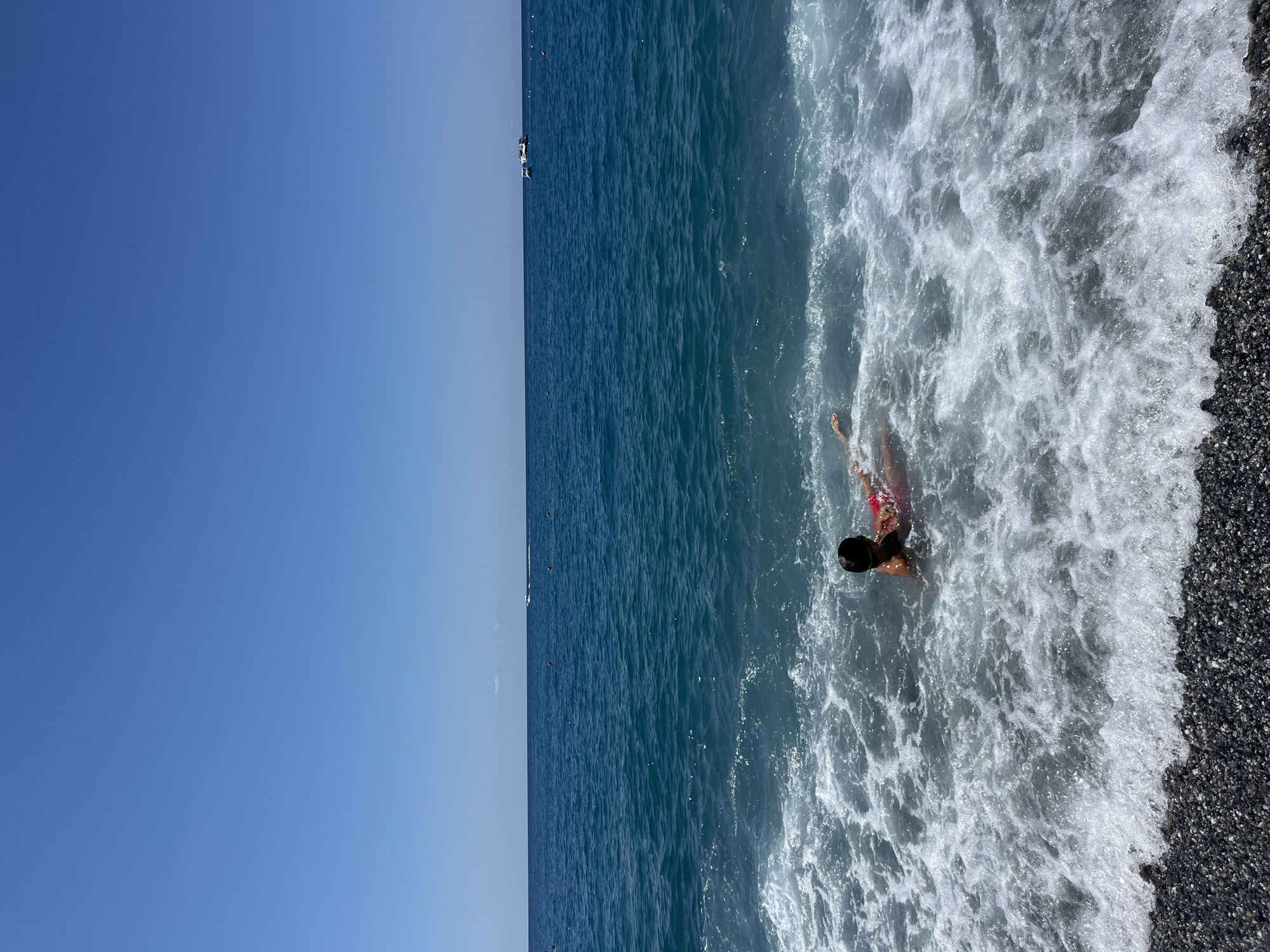 A person wearing a red swimsuit and green goggles is in the shallow water near the shore of the Tyrrhenian Sea in Scalea, Italy. The person is lying on their back in the foamy surf, with their arms outstretched and legs extended. The water is a deep blue, with white waves crashing onto a pebbled beach. In the distance, several boats are visible on the horizon, and the sky is clear and blue. The scene captures a moment of leisure and enjoyment on a sunny day at the beach.