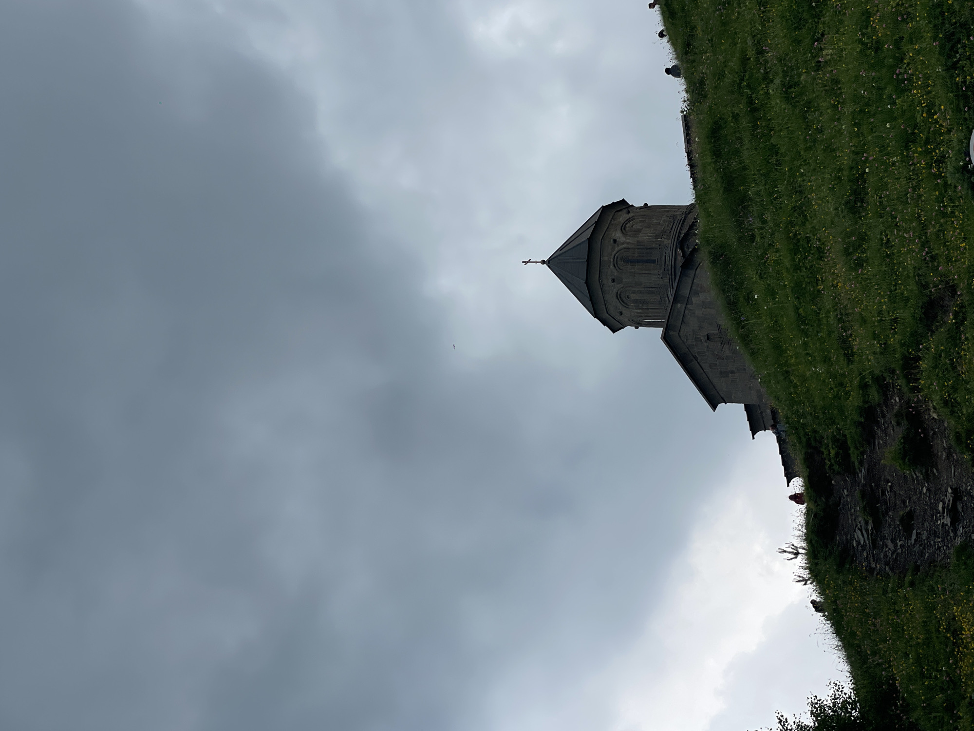 A low-angle, full outdoor shot captures the Gergeti Trinity Church in Stepantsminda, Georgia, under a cloudy sky. The stone church, topped with a dark, conical roof and a cross, is situated on a grassy, sloping hill. Several small figures, presumably tourists, are scattered on the hillside near the church, some of them walking up a rocky path. The hill is covered in lush green grass dotted with wildflowers. The sky is overcast with heavy gray clouds.