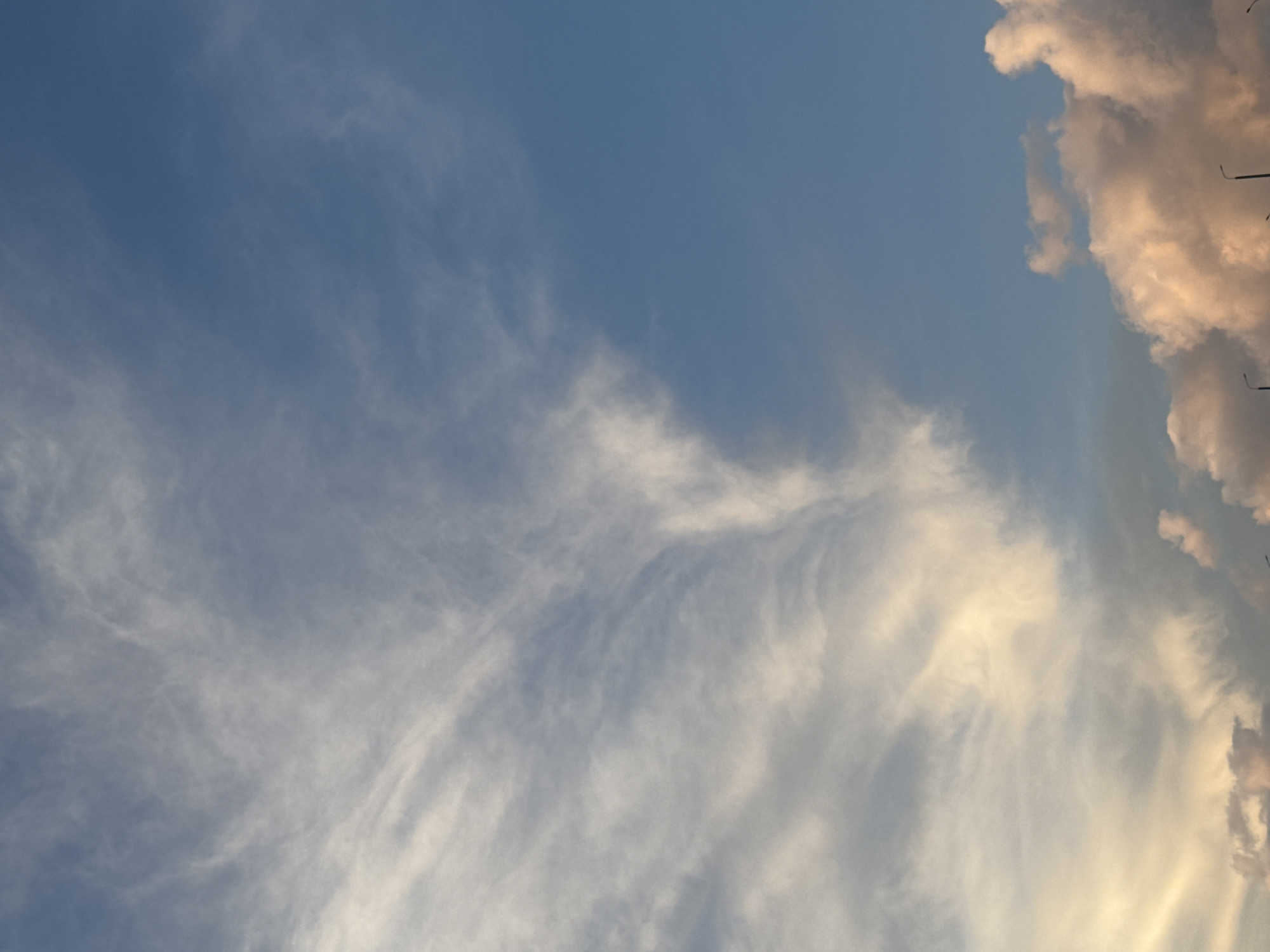 The image depicts a sky in Krasnoyarsk, Russia. The upper right corner contains cumulus clouds with a warm, pinkish hue from the setting sun. The remaining portion of the sky is light blue with cirrus clouds. Utility poles are seen in the upper right corner.