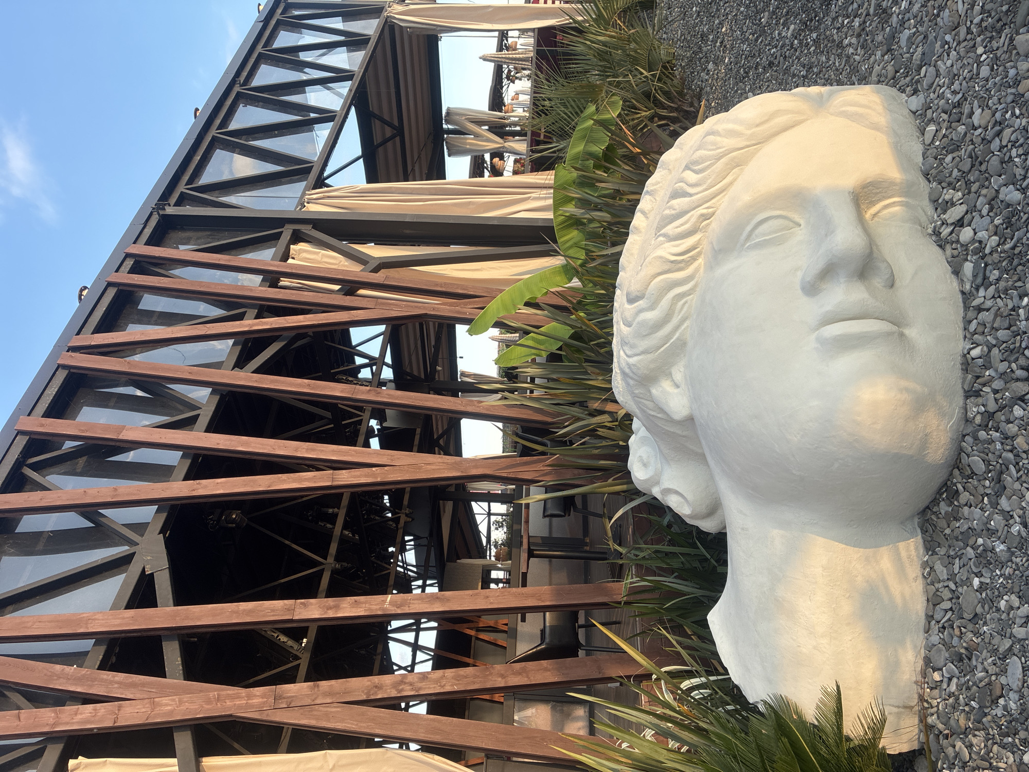 This is an outdoor photograph taken at a beachside restaurant in Sochi, Russia. In the foreground, a large, white sculpture of a reclining human head and shoulders is placed on a bed of grey pebbles. The sculpture is stylized with wavy hair and closed eyes. Behind the sculpture, lush green tropical plants, including palm leaves and banana leaves, are visible.
In the mid-ground, a modern, open-air restaurant structure is visible. It features a dark metal frame supporting a sloping roof with triangular glass panels. Numerous dark wooden beams are diagonally positioned along the facade, creating an interesting geometric pattern. Beige curtains hang from the sides of the structure, partially obscuring the interior. Inside the restaurant, tables and chairs are set up, with some tables appearing to have tablecloths.
The background shows more of the restaurant's open-air seating area, including umbrellas and decorative lights. The sky above is clear and blue with a few wispy clouds, and the setting sun casts a warm, golden light on the scene, highlighting the sculpture and the plants. The overall environment suggests a serene and picturesque location.