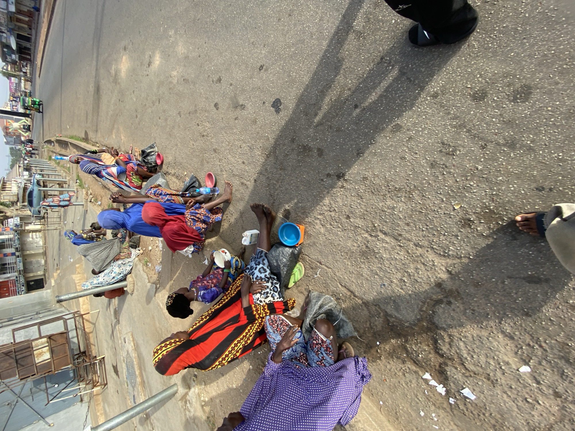 The image depicts several individuals lying on a roadside in Jos, Nigeria. The ground appears to be a paved surface, possibly a sidewalk or edge of a road, with some visible debris. The individuals are lying close together, some covered with blankets or wearing colorful clothing. Some have bowls or containers near them. Shadows indicate the presence of other individuals standing nearby, potentially observing the scene. The background includes buildings, vehicles, and street infrastructure, typical of an urban environment.