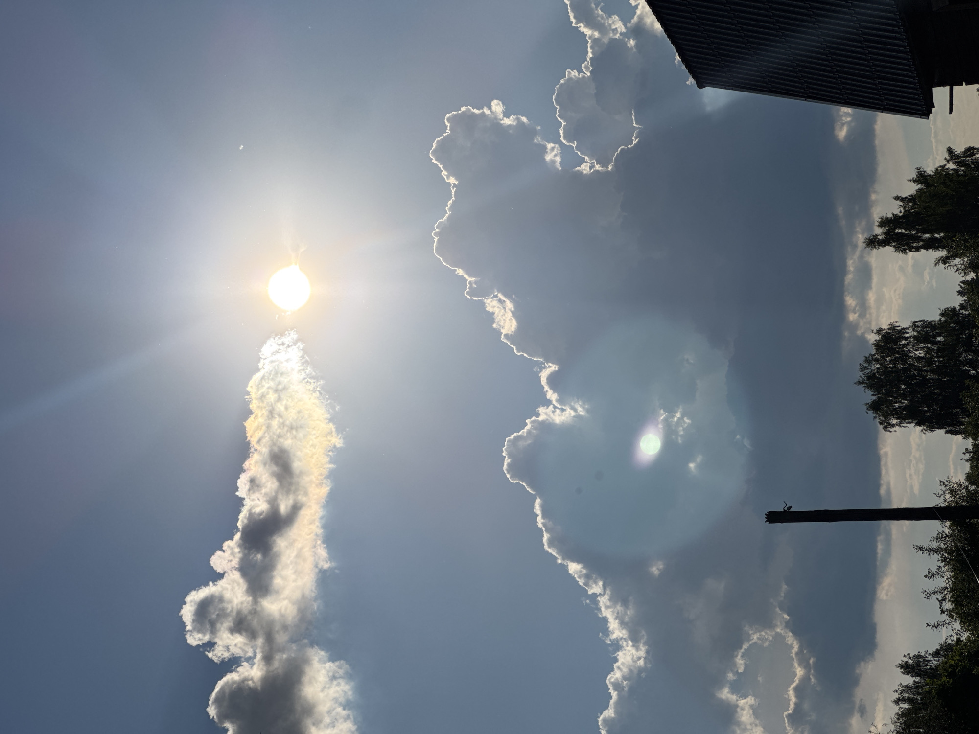 The image depicts a sky filled with dramatic clouds illuminated by the sun. In the upper portion of the frame, the sun is a bright orb, partially obscured by a thin, wispy cloud band that shows iridescence with hints of yellow, orange, and blue. Below this, larger, darker clouds form massive formations with bright, illuminated edges, suggesting they are catching the sunlight from behind. In the lower part of the image, silhouetted against the sky are trees and the dark roof of a building. A single, dark wooden pole stands vertically in the mid-ground. The overall scene is one of a dynamic sky, likely captured in the town of Krivandino, Russia, given the context. There are no people or animals visible in the frame.