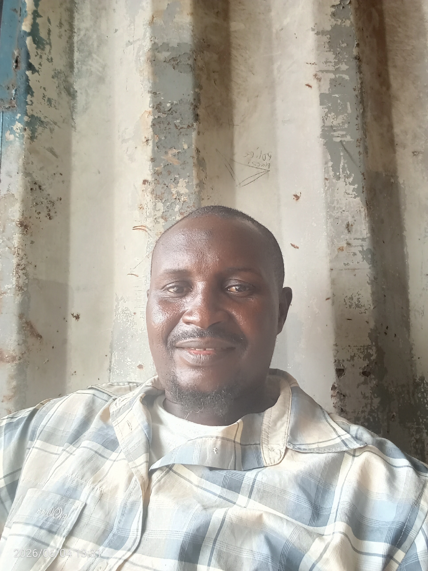 A portrait-style photograph captures an adult Black male, likely in Iddah, Nigeria, smiling faintly and looking directly at the camera. He has short dark hair and a short, dark beard or goatee. He wears a light-colored plaid button-up shirt, with checks in shades of white, blue, and beige, over a white undershirt.
The background is a weathered, corrugated metal wall, possibly part of a shipping container or an industrial building. The wall shows extensive peeling paint in light grey, white, and faint blue tones, revealing rust and general discoloration. Faint, hand-drawn or scratched text and symbols are visible on the wall, including what appears to be "Dilly" and "2019" with a triangular design to the upper left of the man's head.
A digital timestamp "2026/06/04 13:31" is visible in the bottom-left corner of the image. The lighting suggests it is daytime. The man appears relaxed and approachable.