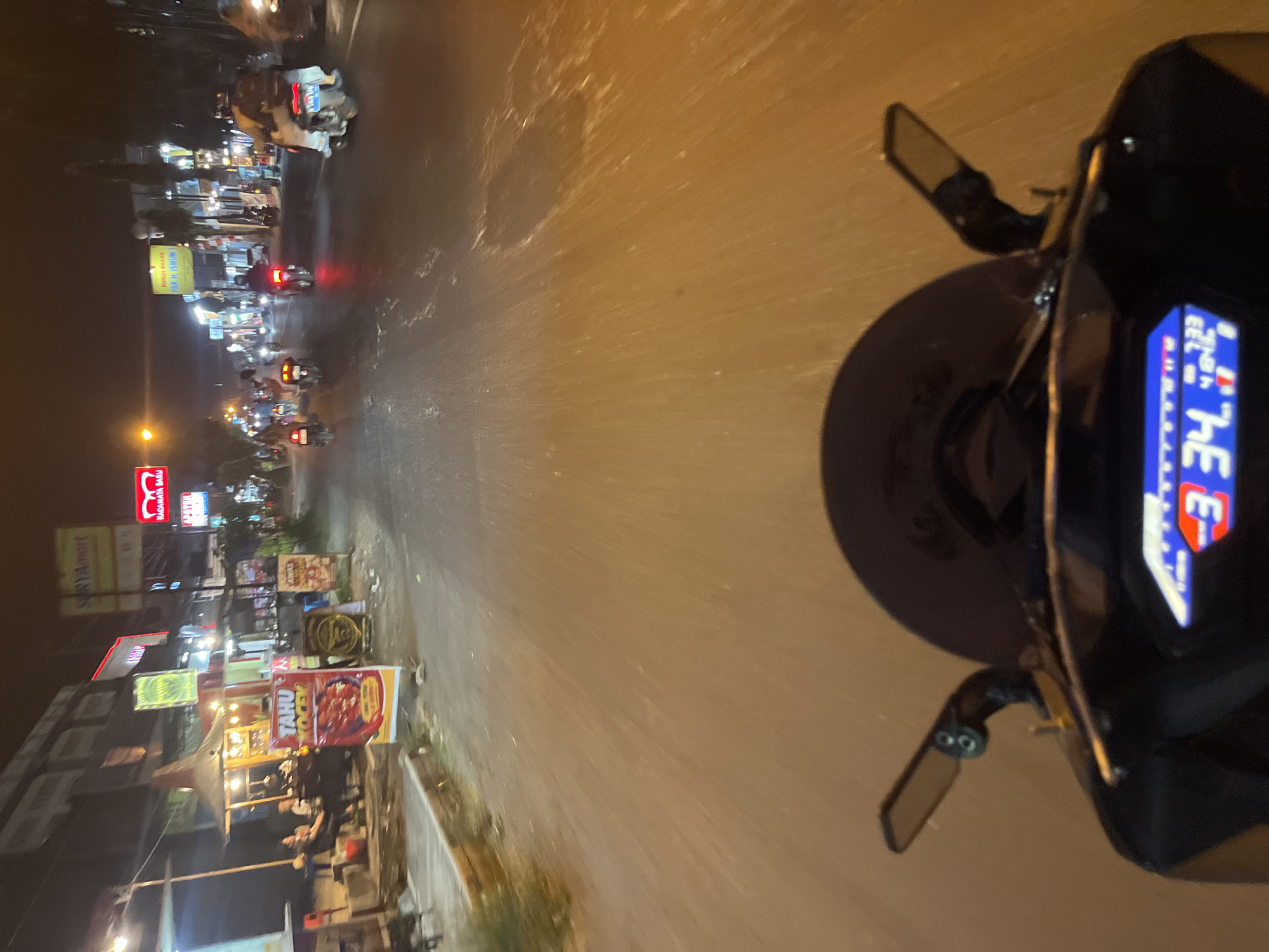 The image depicts a nighttime view from the perspective of a motorcycle rider traveling along a street in Kalinyamatan, Indonesia. The road is paved and wet, reflecting the lights from surrounding businesses. Several motorcycles are visible on the road, some with riders. Along the left side of the street, there are buildings with illuminated signs and storefronts, including what appears to be a food stall with a prominent sign advertising "TAHU VOCEK." On the right side of the street, more buildings and illuminated signs are present. The rider's motorcycle dashboard, displaying a speed of 34 km/h, is in the foreground, along with a rearview mirror. The overall scene is characterized by artificial lighting, traffic, and commercial establishments typical of a town at night.