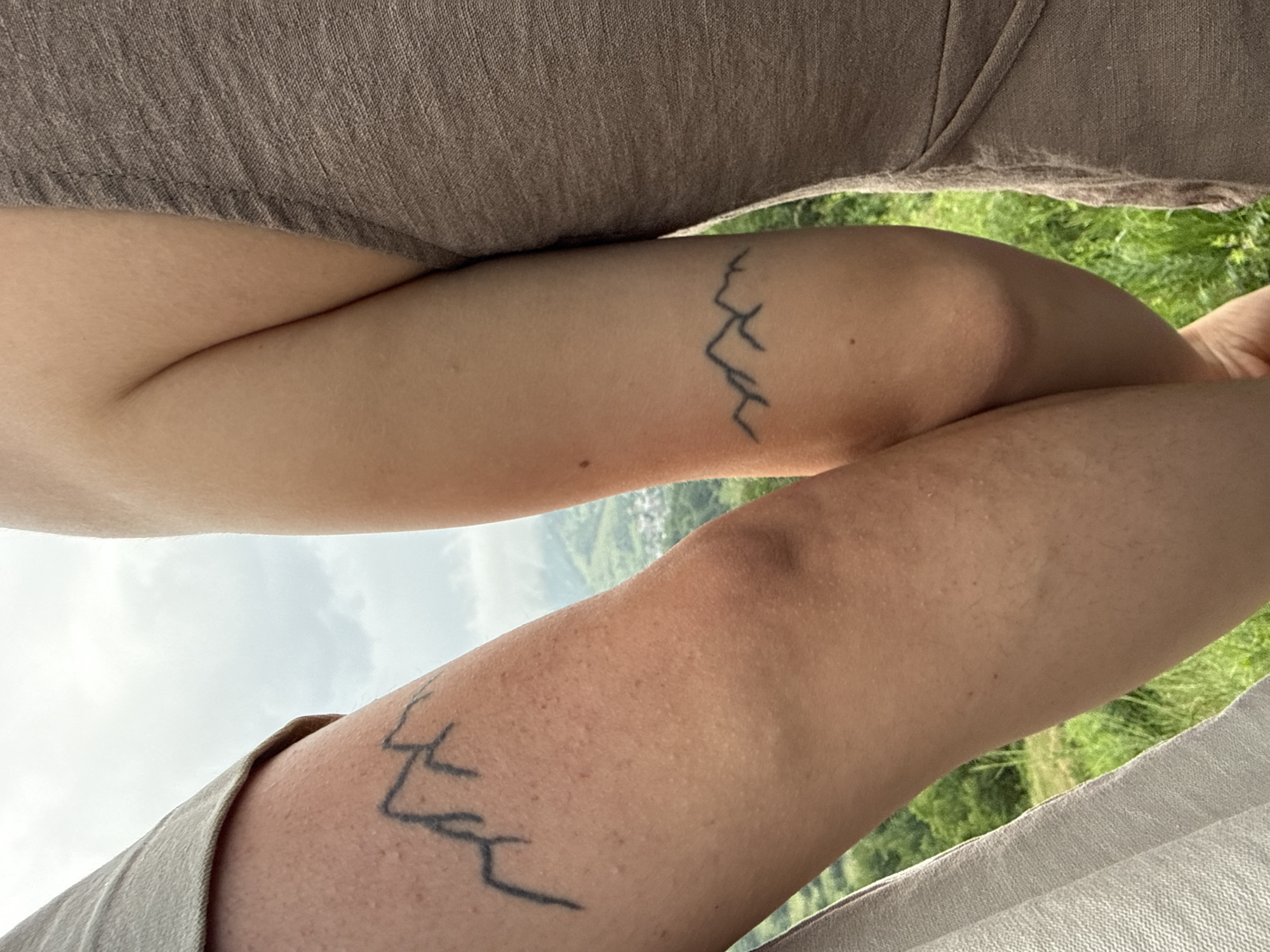 The image presents a close-up of two human forearms, held in a close, seemingly intertwined position, against a backdrop of a verdant, hilly landscape under an overcast sky. The arm on the left, displaying a minimalist black outline tattoo of a three-peaked mountain range on its upper forearm, is partially covered by a light, finely woven, possibly beige or off-white fabric garment. The arm on the right features a similar, horizontally elongated black outline tattoo of a multi-peaked mountain range on its lower forearm, and is partially clad in a brown, textured fabric garment with a subtle draped or folded detail. Both arms exhibit visible skin texture and minor freckling. In the mid-ground and background, lush green rolling hills and valleys unfold, with a small cluster of buildings, indicative of a village or town, visible nestled among the trees. The sky above is largely cloudy, with diffused light. This scene is situated in the city of Ardino, Bulgaria.