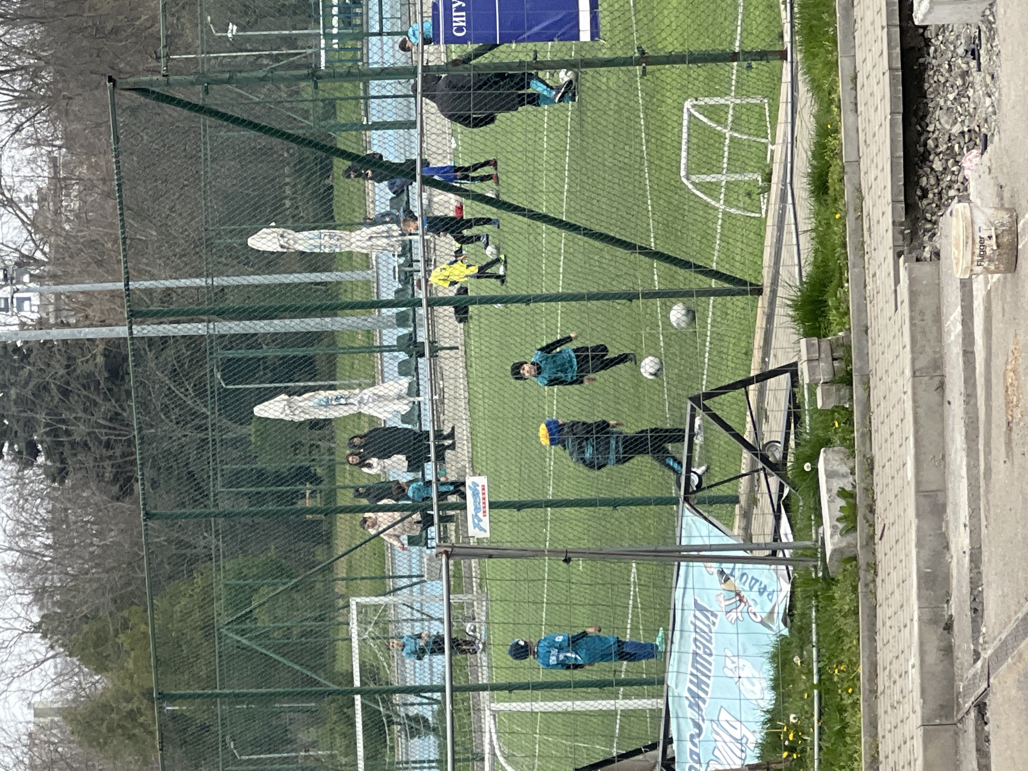 A youth soccer game is being played on an artificial turf field enclosed by a chain-link fence. Several young players in different colored uniforms are on the field. Adults are standing along the sidelines, watching the game. Two soccer goals are visible, one at each end of the field. The game is occurring in Burgas, Bulgaria, based on the Cyrillic writing on a banner on the fence. The location is adjacent to a sidewalk or paved area with some construction materials. A few trees are visible beyond the fence.