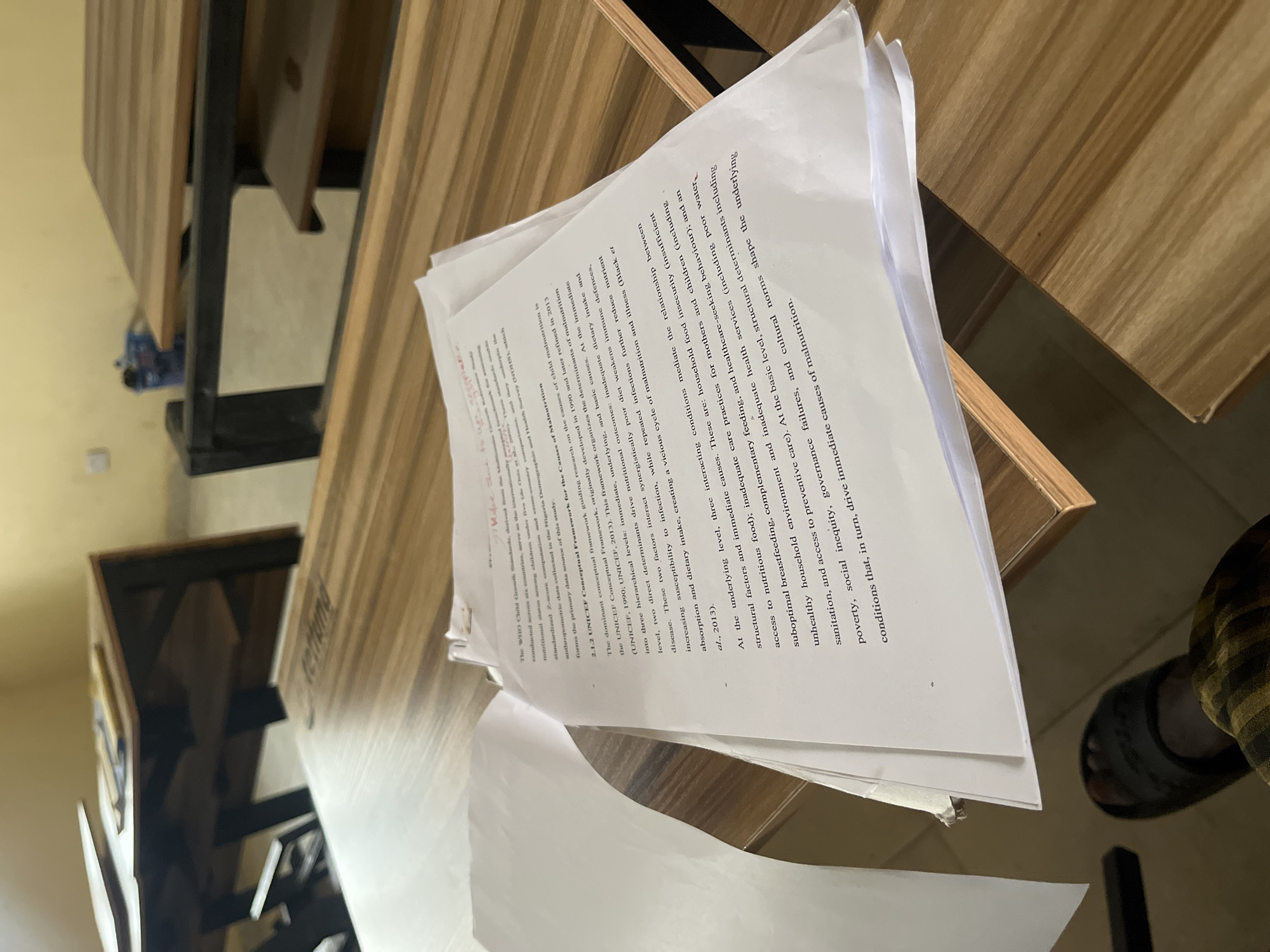 The image depicts a pile of papers on a wooden surface, possibly a table or desk. The papers appear to contain text, with headings like "UNICEF Conceptual Framework for the Causes of Malnutrition" and references to nutritional status in children, mentioning the Nigeria Demographic and Health Survey (NDHS). The text discusses various levels of determinants for malnutrition (immediate, underlying, and basic causes). The wooden surface has a light, wood-grain pattern. Part of a foot wearing a slipper is visible in the lower right corner. The image seems to be taken indoors. There is no indication of the location being in Baganakwo, Nigeria, apart from the reference to the Nigeria Demographic and Health Survey (NDHS).