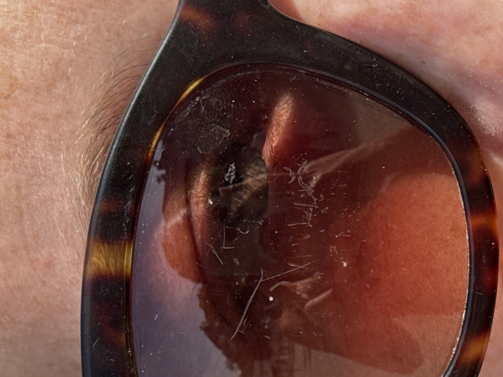 A close-up view shows part of a person's face, including skin with freckles and a light-colored eyebrow, next to a pair of sunglasses. The sunglasses have a brown tortoiseshell pattern frame, and the lens is dirty with scratches, dust, and what appears to be small fibers. The reflection on the lens of the sunglasses gives a distorted view of the skin and facial features on the person's face. The background is indistinct. There is no information suggesting that this image was taken in Paris, France.