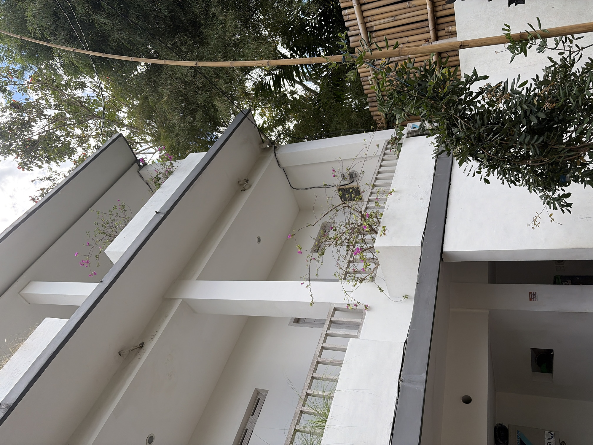 The image shows a multi-story building with white walls and dark gray trim, located in Mengwi, Indonesia. The building has balconies with wooden railings. Pink bougainvillea flowers are visible on the balconies. In the foreground, there is a dense green bush next to a tall, slender bamboo pole. In the background, lush green trees and bamboo foliage can be seen, along with a bamboo fence. The sky is partially visible, with some clouds. Electrical wires are strung across the scene. The lower part of the building reveals an open-air covered area with part of an air conditioning unit visible on the left.
