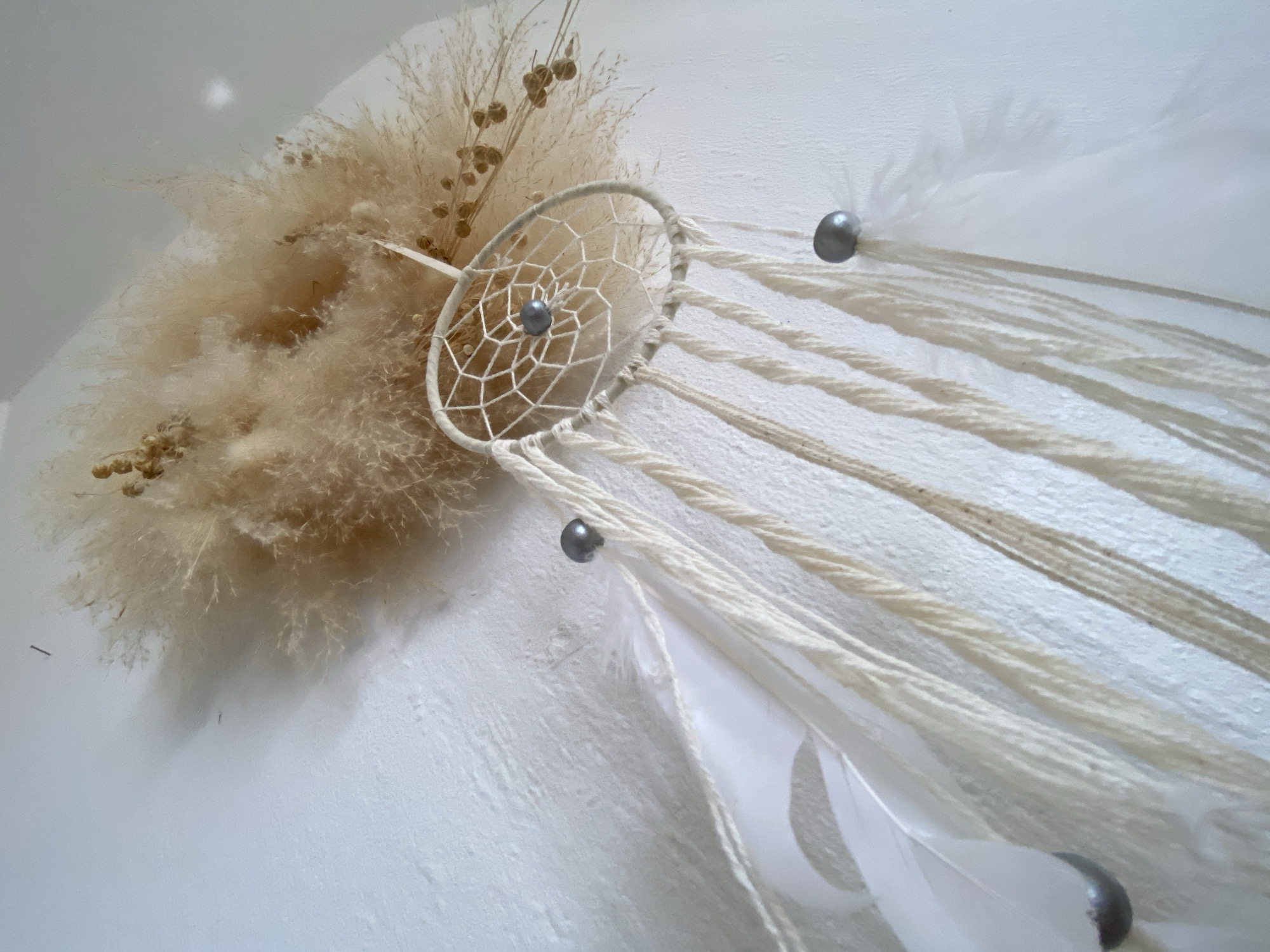A dreamcatcher is hanging on a textured white wall. The dreamcatcher has a white hoop with a web pattern, and a single grey bead in the center. Dangling from the hoop are strands of cream-colored yarn and white feathers. There are also two grey beads on the strands. Behind the dreamcatcher, a bundle of dried beige grasses and some small seed pods are attached to the wall. The lighting suggests this is an indoor setting. The image does not contain any information that would indicate its location in Bad Sachsa, Germany.