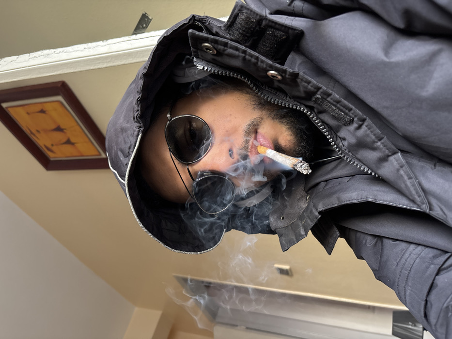 The image displays a close-up, eye-level view of a single individual in an indoor setting in Tangier, Morocco. The subject, a person with facial hair, occupies the majority of the frame, facing slightly left. They are wearing dark, round-lensed sunglasses and a black hooded jacket with a white stripe visible along the inner edge of the hood. A lit, hand-rolled cigarette is positioned between their lips, emitting a substantial amount of white smoke that partially obscures the lower portion of their face and drifts towards the left side of the frame. The background consists of a plain, light beige-colored wall. In the upper right quadrant, a framed piece of artwork hangs on the wall, depicting three abstract, stylized figures with yellow or orange bodies and dark heads, set against a darker background within a dark reddish-brown frame. To the far right, a vertical white architectural element, likely a door frame, is partially visible. To the far left, another light-colored vertical structure, possibly a wall or furniture, is partially visible. No other individuals or direct interactions are present in the scene.