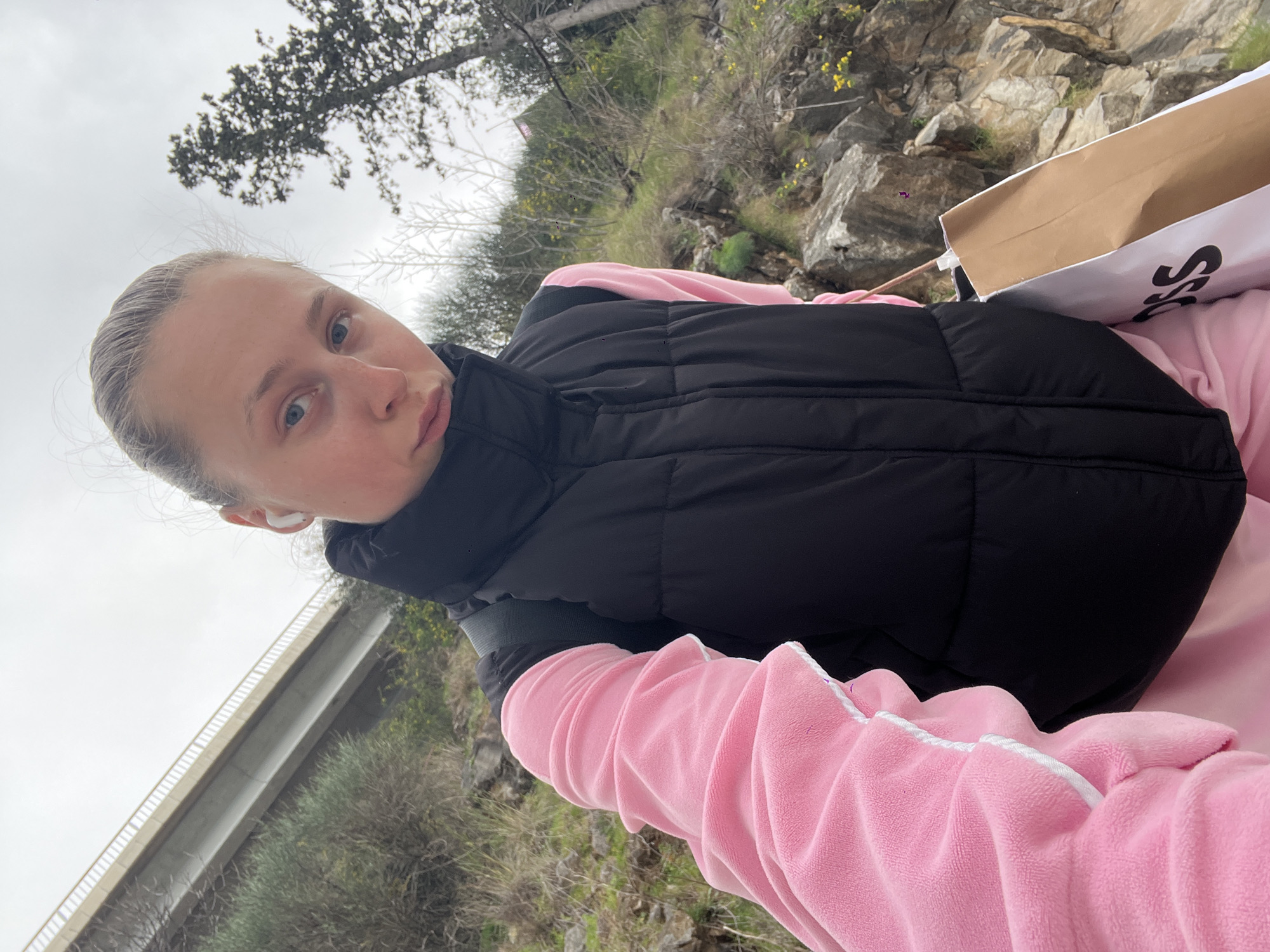A fair-skinned woman is holding a camera up to take a selfie. She has light blue eyes and her light-colored hair is pulled back. She is wearing a pink velour tracksuit and a black puffer vest. She has a wireless earbud in her ear. She is holding a brown and white shopping bag. Behind her are rocks and greenery. A concrete bridge can be seen in the background. The location is in Budva, Montenegro.