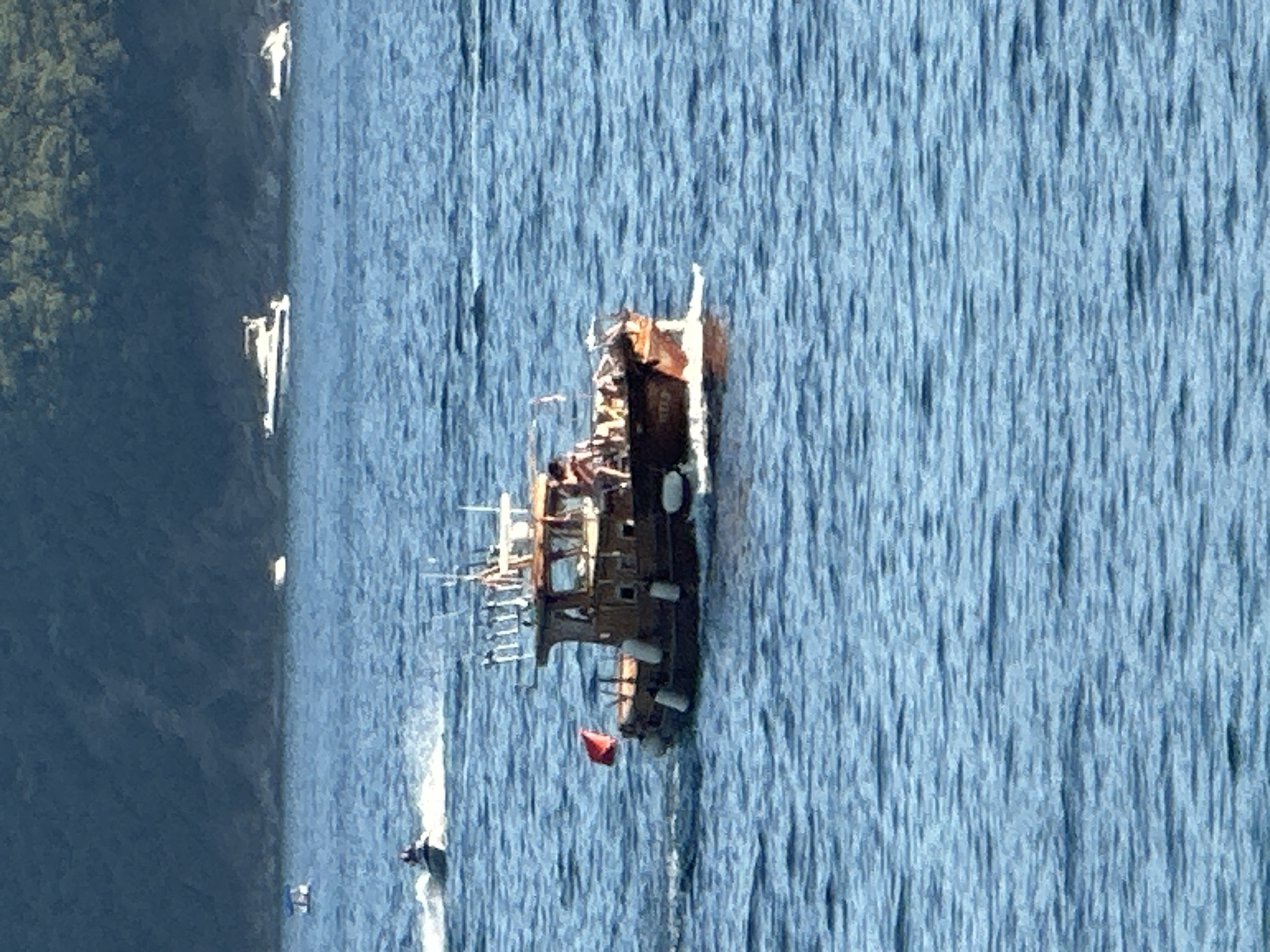 The image depicts a serene body of water, likely a bay or sea, with a wooded hillside in the background. Several boats are visible. In the foreground, a wooden motorboat with a cabin is sailing, displaying a red flag on its port side and a nameplate that appears to read "SUNBEAM". The boat has several people on board and fishing rods set up. To the left of the motorboat, a jet ski is in motion, leaving a white wake. Further in the background, two white motorboats are anchored or cruising, and another small vessel with a blue canopy is also present. The water is calm with gentle ripples, and the sky appears to be clear. The overall impression is a tranquil day on the water, possibly in a scenic coastal area. Given the information about Budva, Montenegro, this scene could be taking place in the Adriatic Sea, near the coast of Budva, known for its beautiful beaches and clear waters.