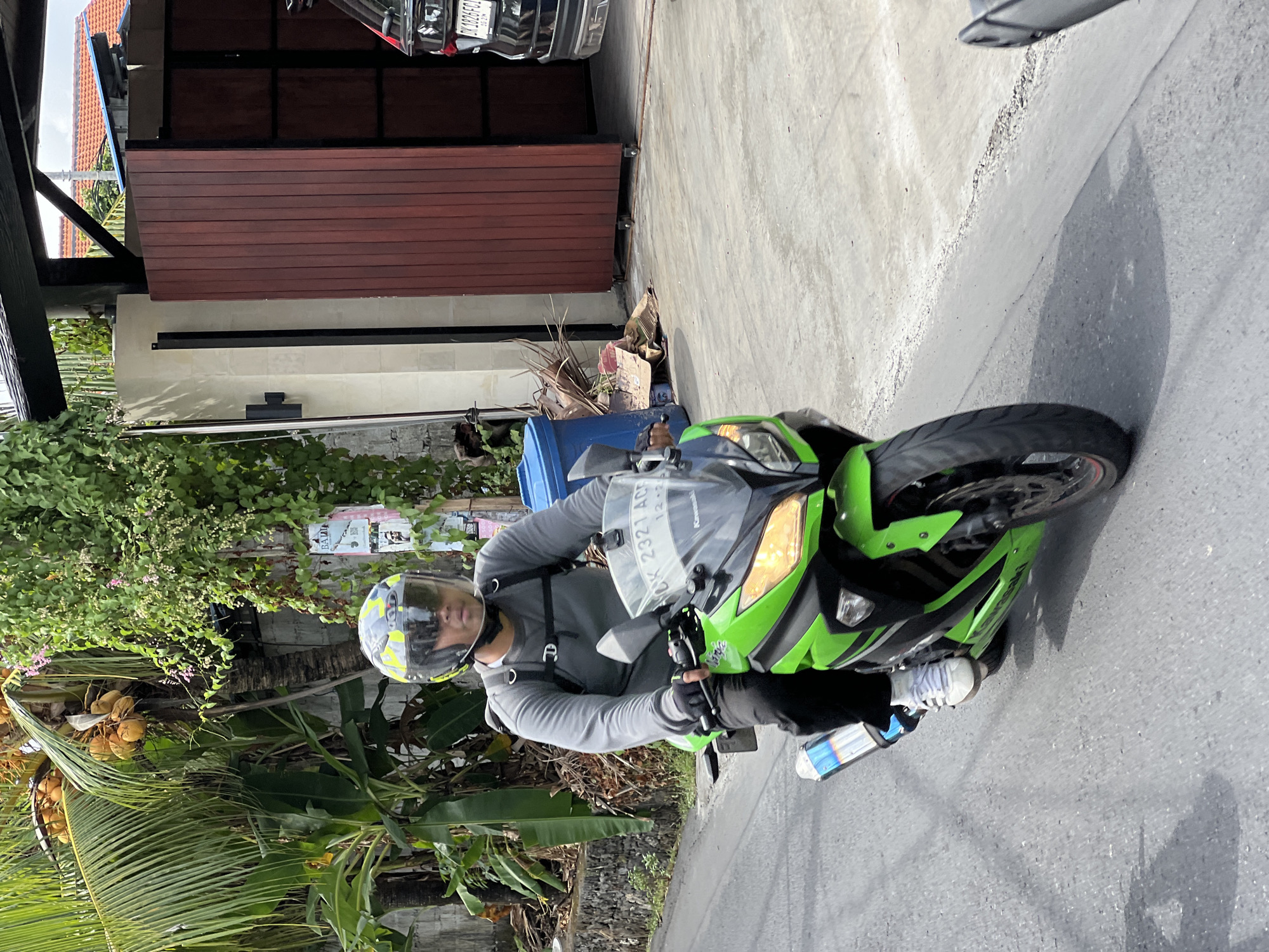 A medium shot captures a person riding a bright green Kawasaki Ninja motorcycle down a paved street in North Kuta, Indonesia. The rider, wearing a silver and yellow patterned helmet with a clear visor, dark pants, white sneakers, and a gray long-sleeved shirt, is looking ahead. They have a black backpack with silver accents secured to their shoulders. The motorcycle features black tires, silver rims with red accents, and a prominent exhaust pipe. In the background, lush green vegetation, including palm trees with coconuts, covers a concrete wall. To the right of the frame, a large wooden gate with vertical planks stands open, revealing a portion of a dark-colored SUV with a license plate that reads "DK 1826 FC1". A blue trash bin and piles of dried leaves and debris are visible near the gate. The ground is a mix of asphalt and concrete. The lighting suggests it is daytime.