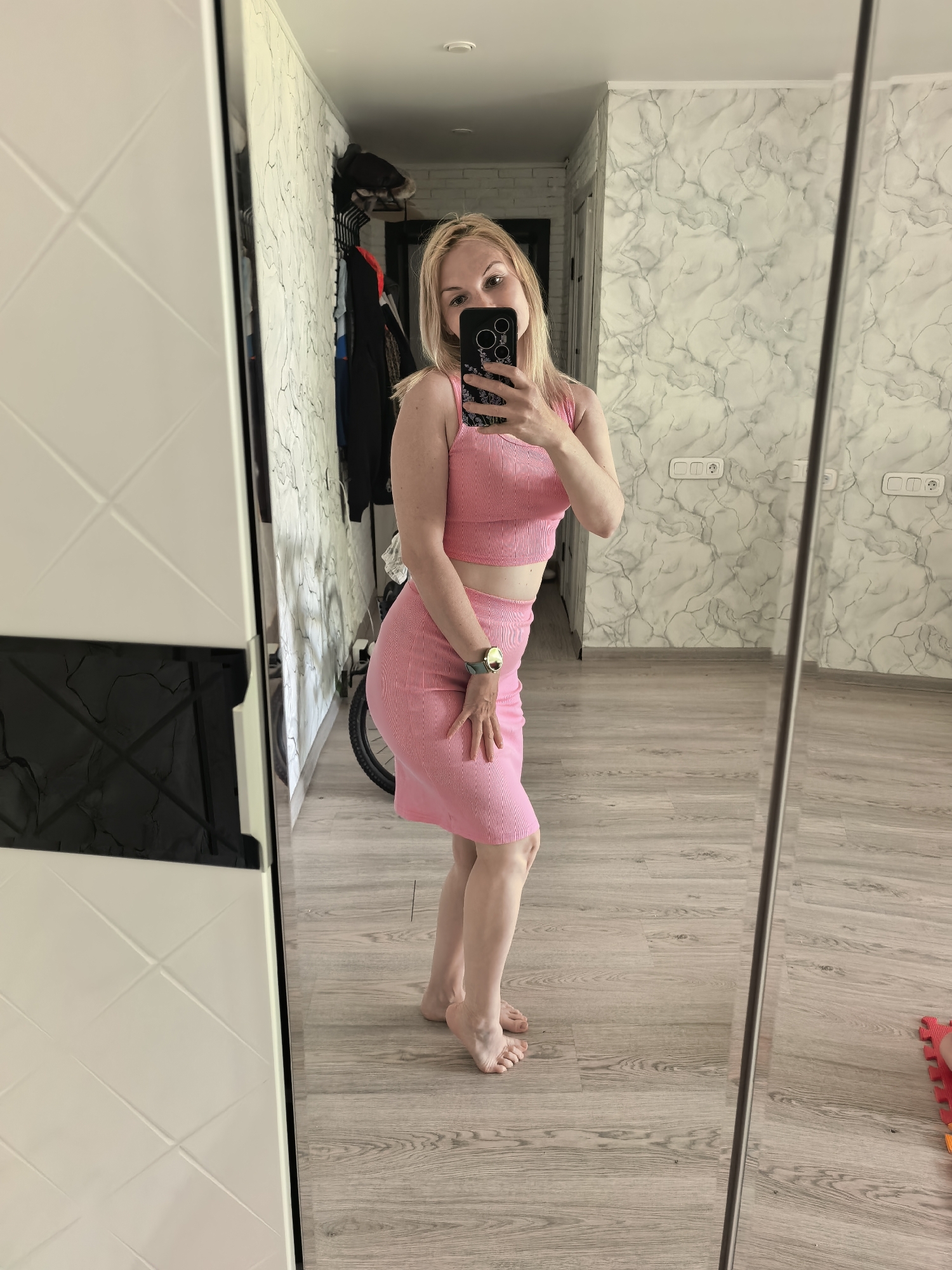 A full-length interior shot shows a blonde woman with blue eyes posing in a mirror selfie. She is wearing a pink two-piece outfit consisting of a crop top and skirt. A wristwatch is visible on her left wrist. The setting appears to be an apartment or home in Sterlitamak, Russia. The walls are light with a marble-like pattern, and the floor has a wood-grain effect. A wardrobe with both white and black panels is visible to the left, with clothes hanging inside. A bicycle is partially visible in the background. Electrical outlets are visible on the right wall. Red children's play mats are located in the bottom right corner.