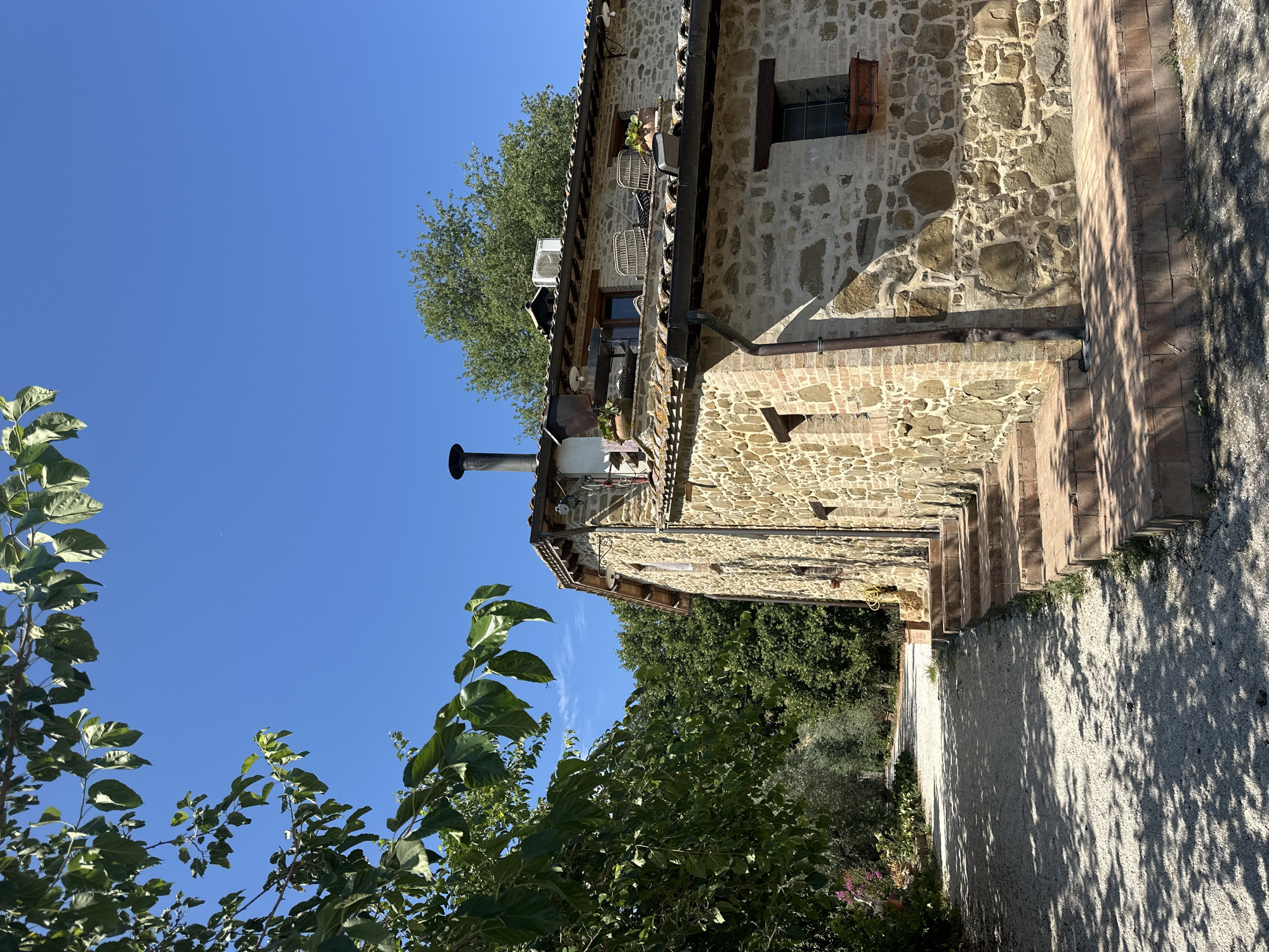 This image captures a sun-drenched stone building with a gravel pathway leading up to it in Cannara, Italy. The building, constructed of large, irregular stones interspersed with brickwork, features a tiled roof. A dark chimney extends from the roofline. Steps made of terracotta bricks ascend to the entrance. Green foliage from trees, casting dappled shadows, frames the scene. The sky is a clear, bright blue. No people are visible in the image. The environment appears to be rural or semi-rural, with mature trees and natural landscaping.