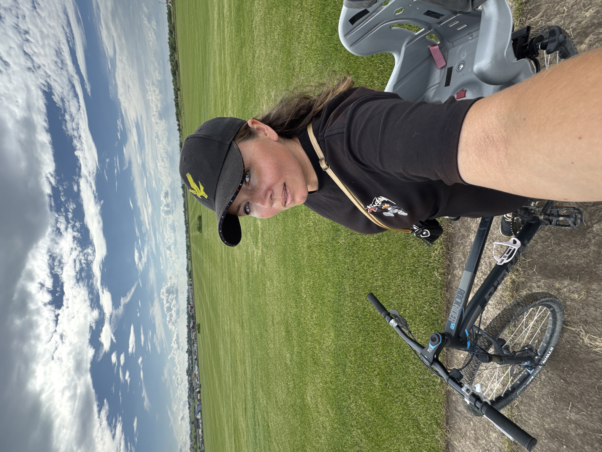 In Kempelevo, Russia, a woman wearing a black baseball cap with a yellow emblem and a black hoodie is taking a selfie while sitting on a bicycle. The bicycle has a child seat attached to the rear. She is looking towards the camera with a neutral expression. The background consists of a vast, well-maintained green field with a few scattered trees and a residential area in the distance. The sky is partly cloudy with a mix of grey and white clouds against a blue sky. The woman is positioned on a dirt path at the edge of the field.