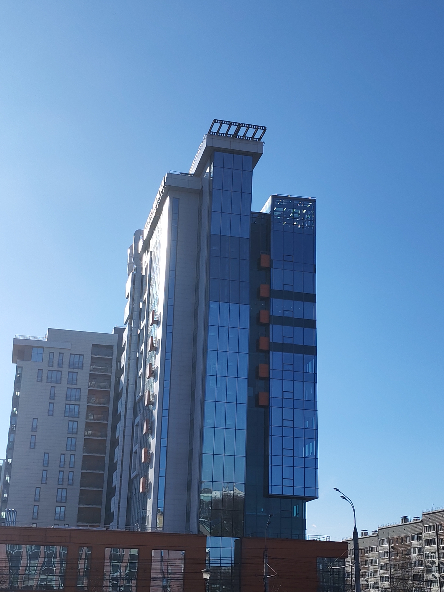 The image shows a tall, modern office building in Izhevsk, Russia, under a clear blue sky. The building is primarily glass and steel, with a smaller building on the left and another building on the right. The reflection of buildings can be seen in the glass of the office building. A streetlight is visible on the bottom right side of the image.